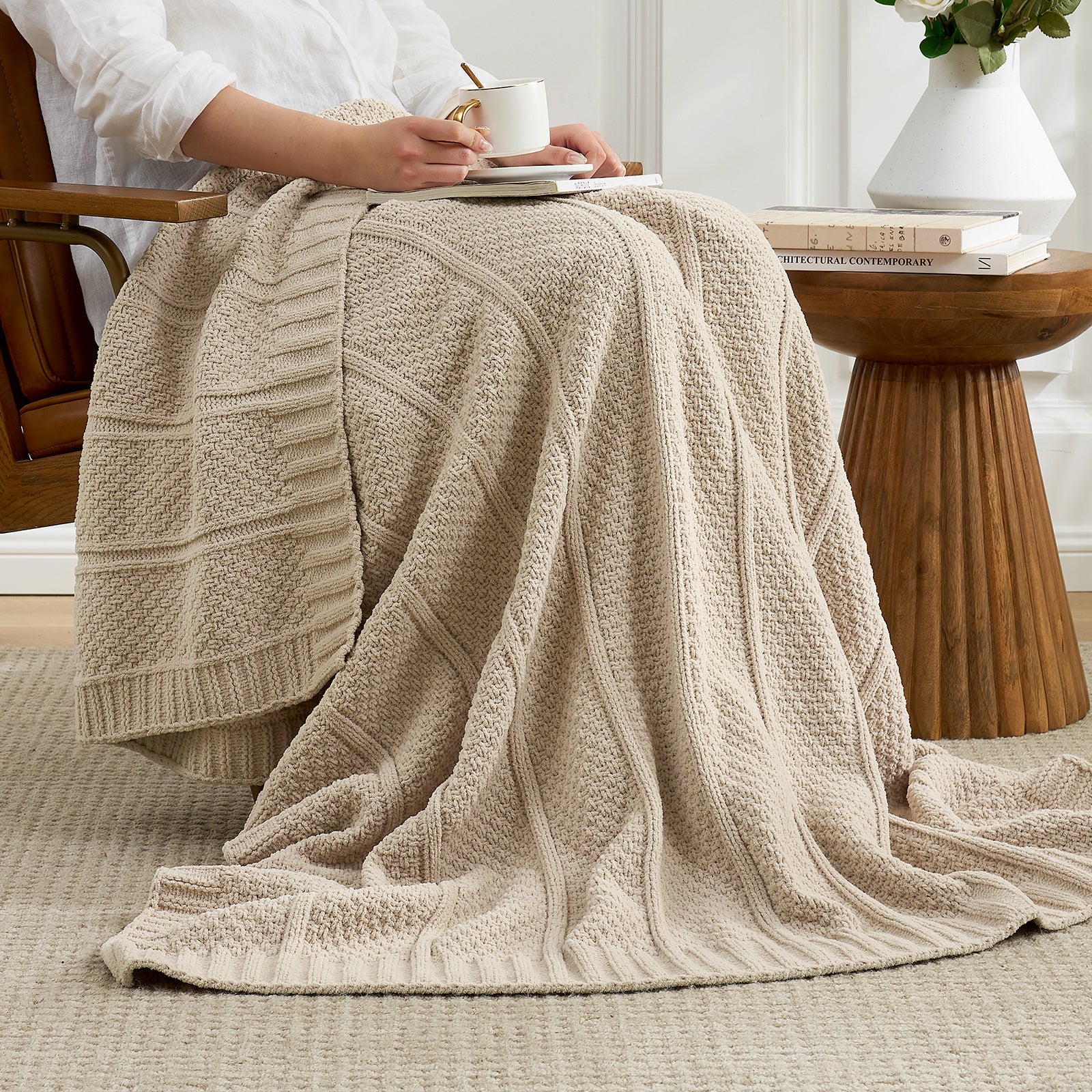 Woman sitting on a chair with a beige knitted blanket draped over her lap, holding a cup. #color_Beige