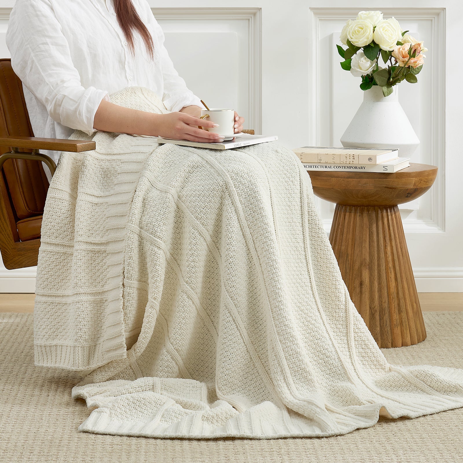 Woman sitting on a chair with an ivory knit blanket draped over her legs, holding a cup. #color_Ivory