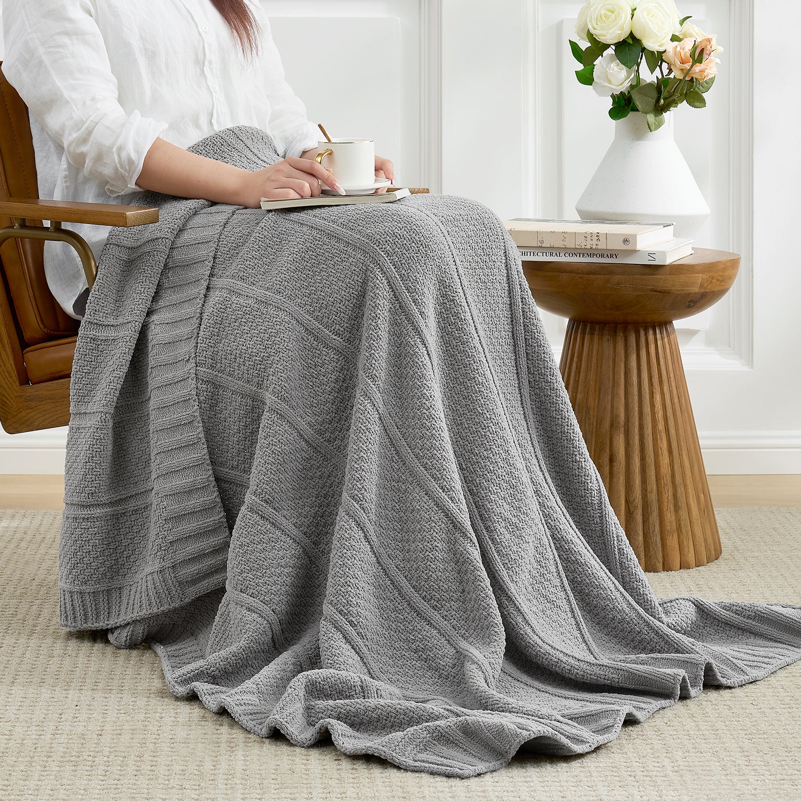 Woman sitting on a chair with a gray blanket draped over her, holding a book and cup. #color_Gray