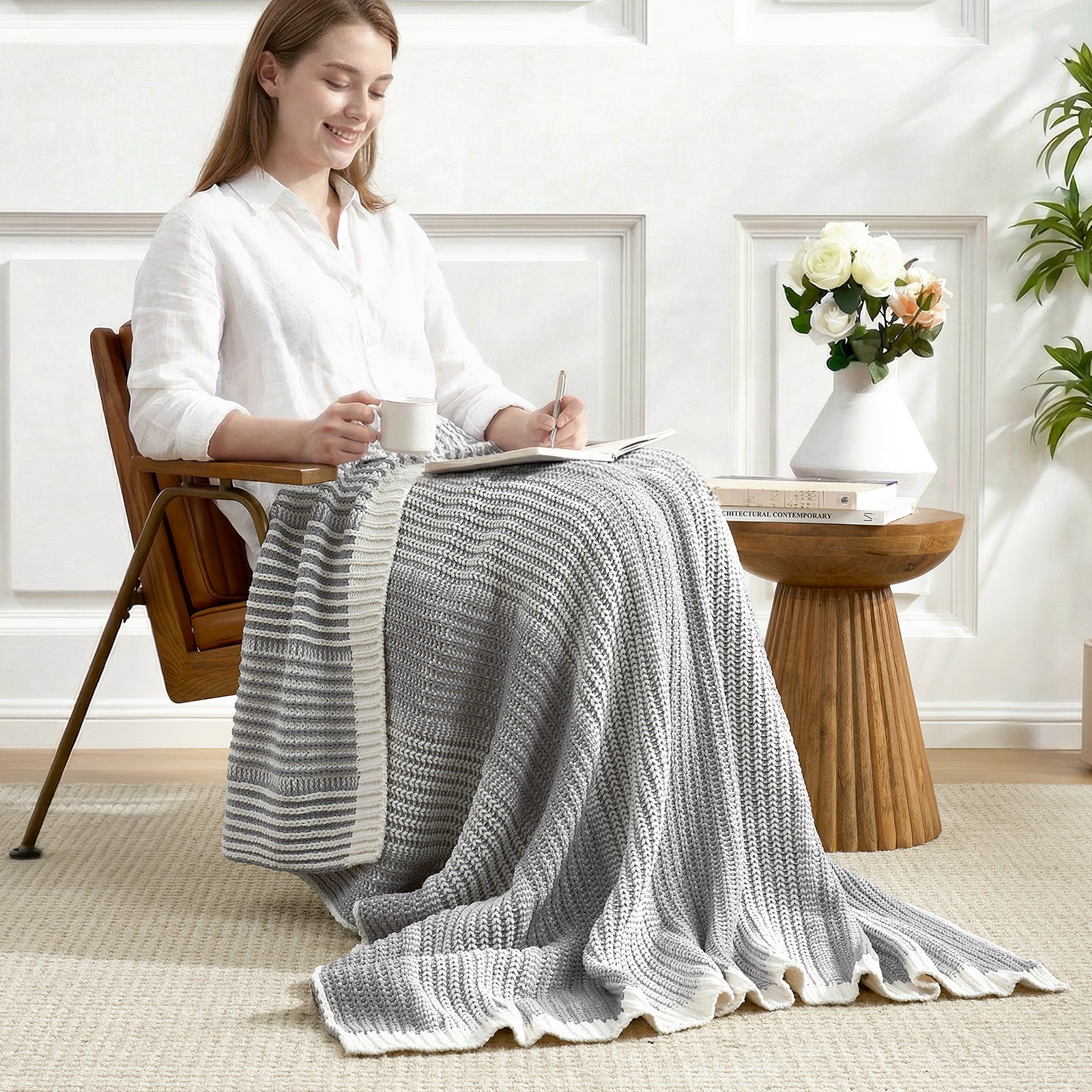 Woman sitting on a chair with a grey knit blanket draped over her lap, holding a cup and pen, in a bright room. #color_Two-tone Grey
