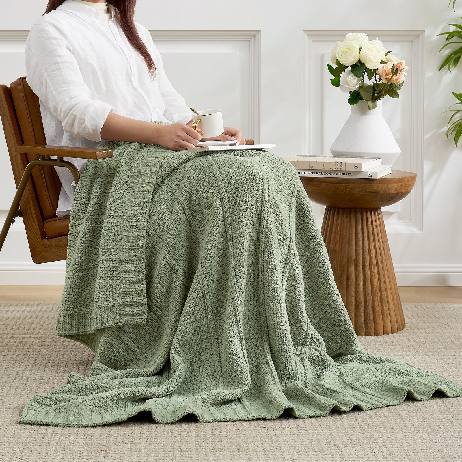 Person sitting in a chair with a green knitted blanket draped over their lap, in a cozy room. #color_Sage Green