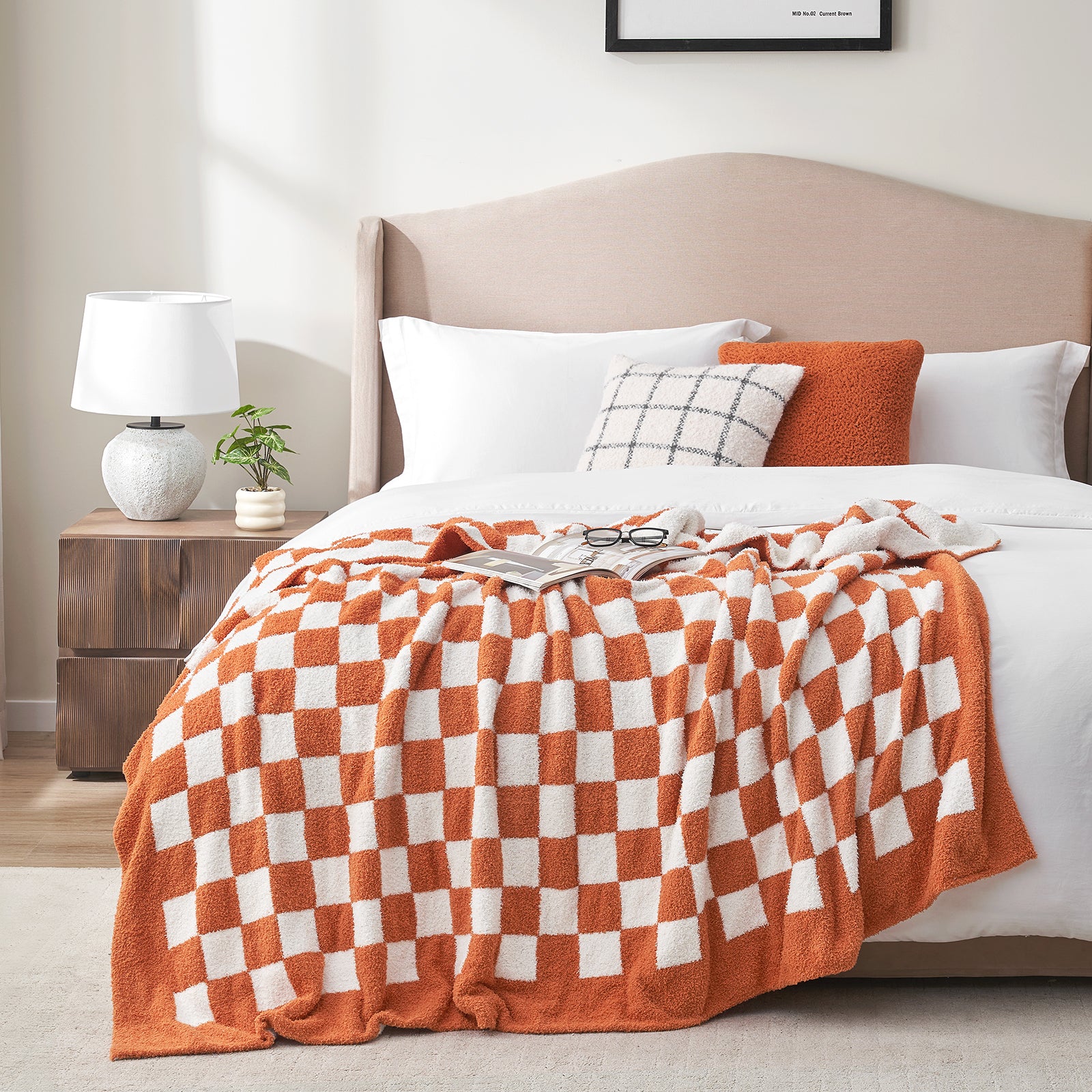 Bedroom with a bed featuring an orange and white checkered blanket, pillows, and a lamp on a nightstand. #color_Checkered Burnt Orange