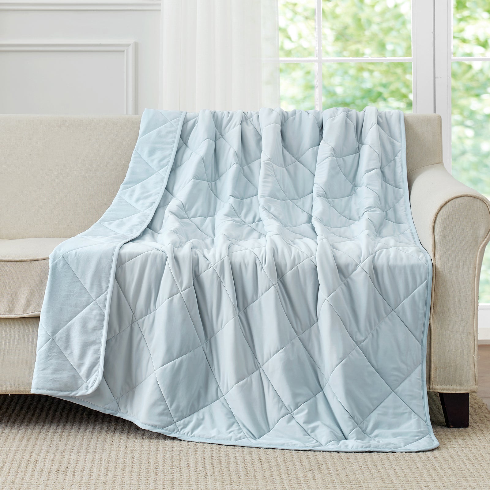 Blue cooling blanket draped over a beige sofa in a room with large windows.#color_Blue