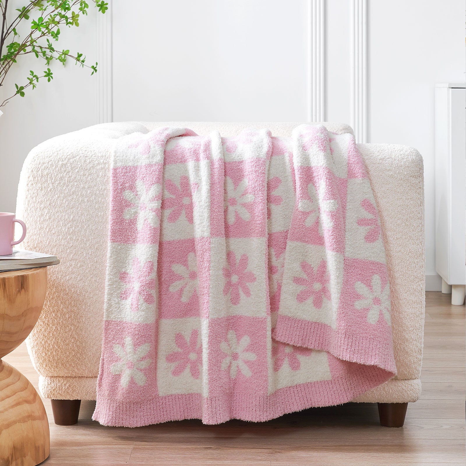 Pink and white floral-patterned blanket draped over a beige ottoman in a room with a wooden table and plant. #color_Baby Pink