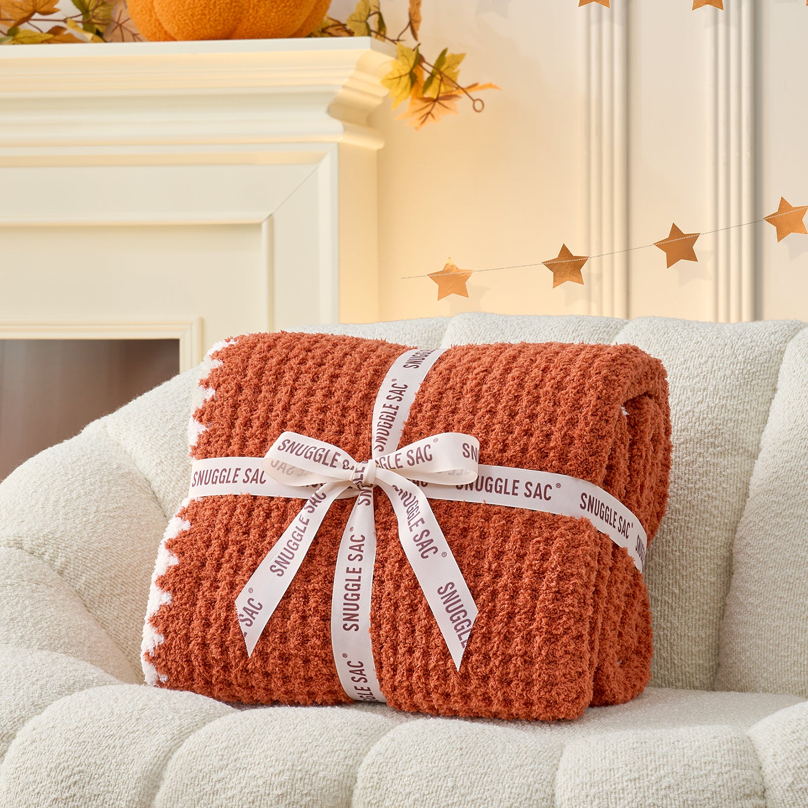 Orange blanket with a white ribbon on a beige couch, with a fireplace and decorative elements in the background. #color_Orange