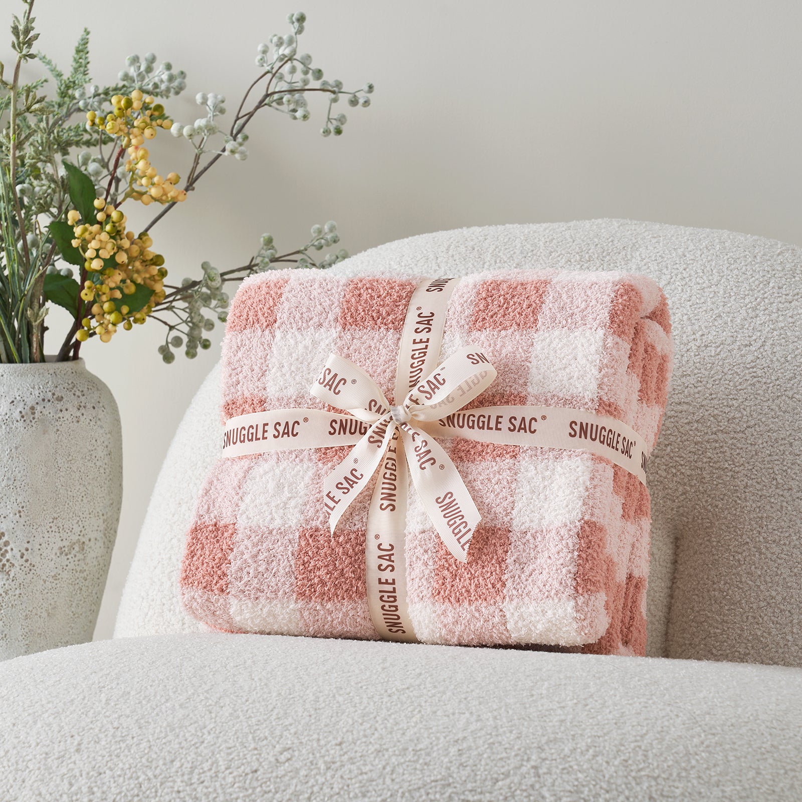 Folded checkered blanket with 'Snuggle Sac' branding on a gray surface, accompanied by decorative plants. #color_Plaid Pink