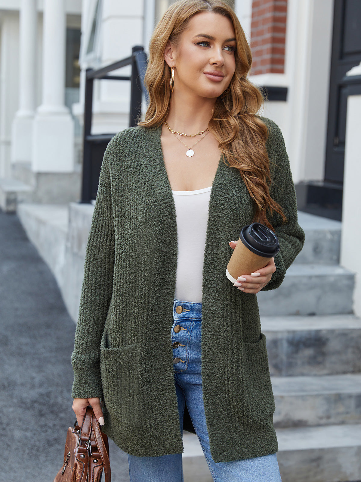 Woman wearing a green cardigan, white top, and blue jeans, holding a coffee cup and a handbag, standing on steps.#color_Army Green