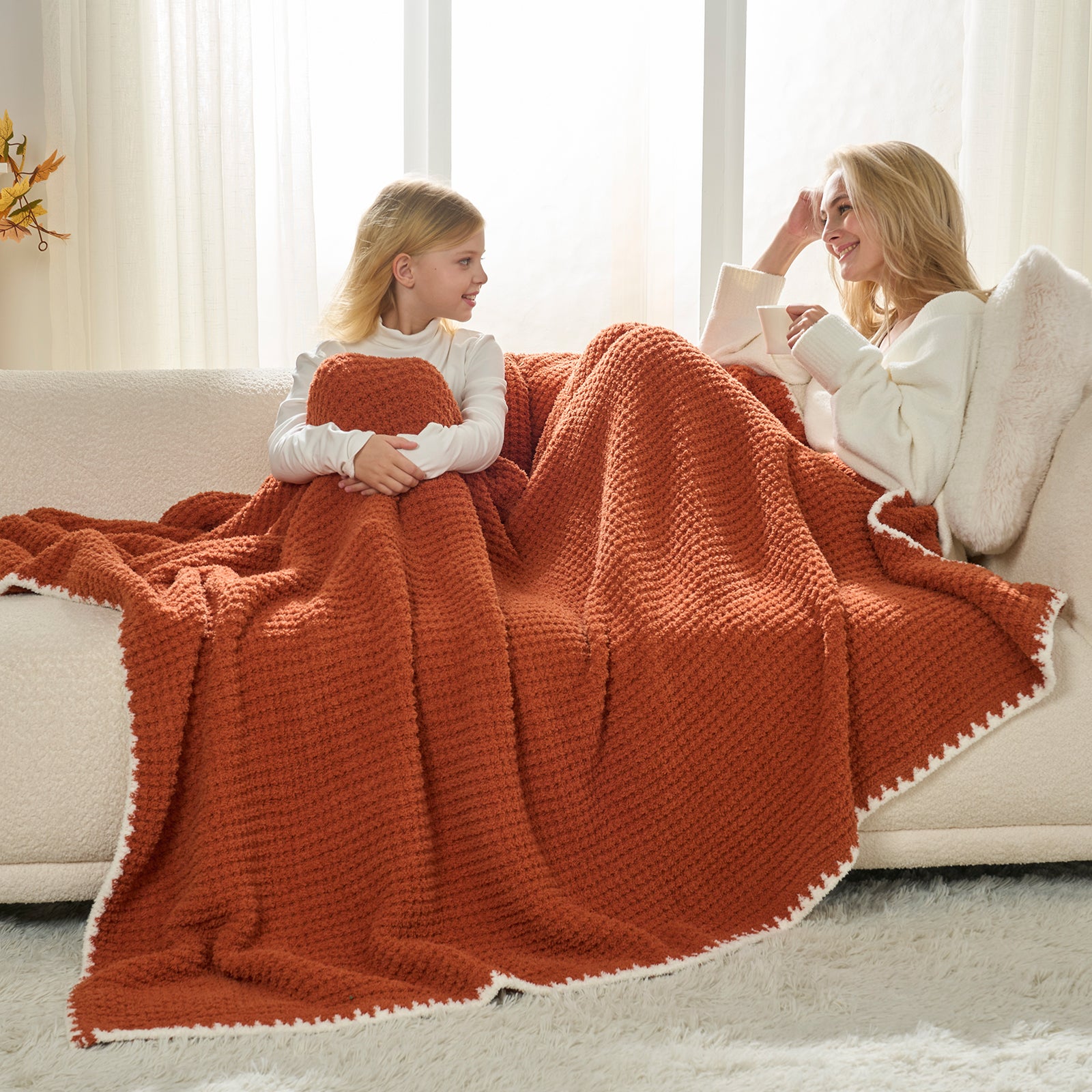 Two people sitting on a couch under a large orange blanket in a bright room. #color_Orange