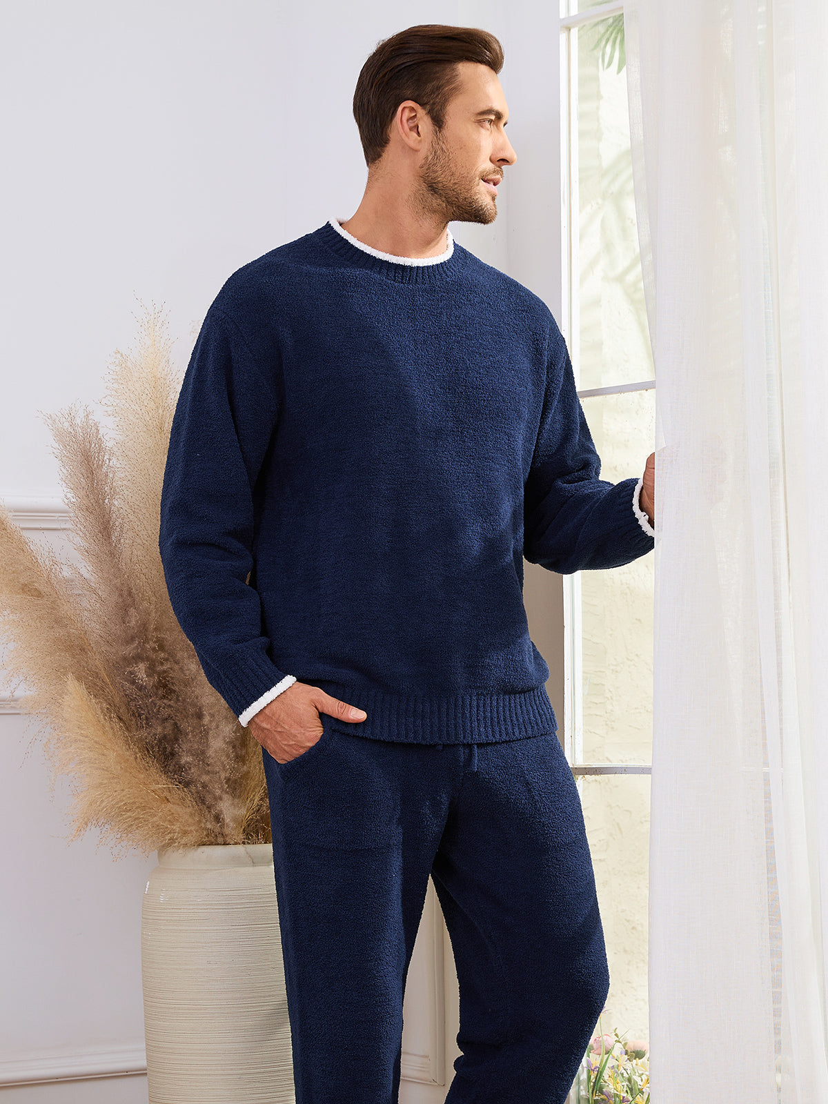 Man wearing a navy blue sweater and pants standing in a room with a vase of dried plants. #color_Navy