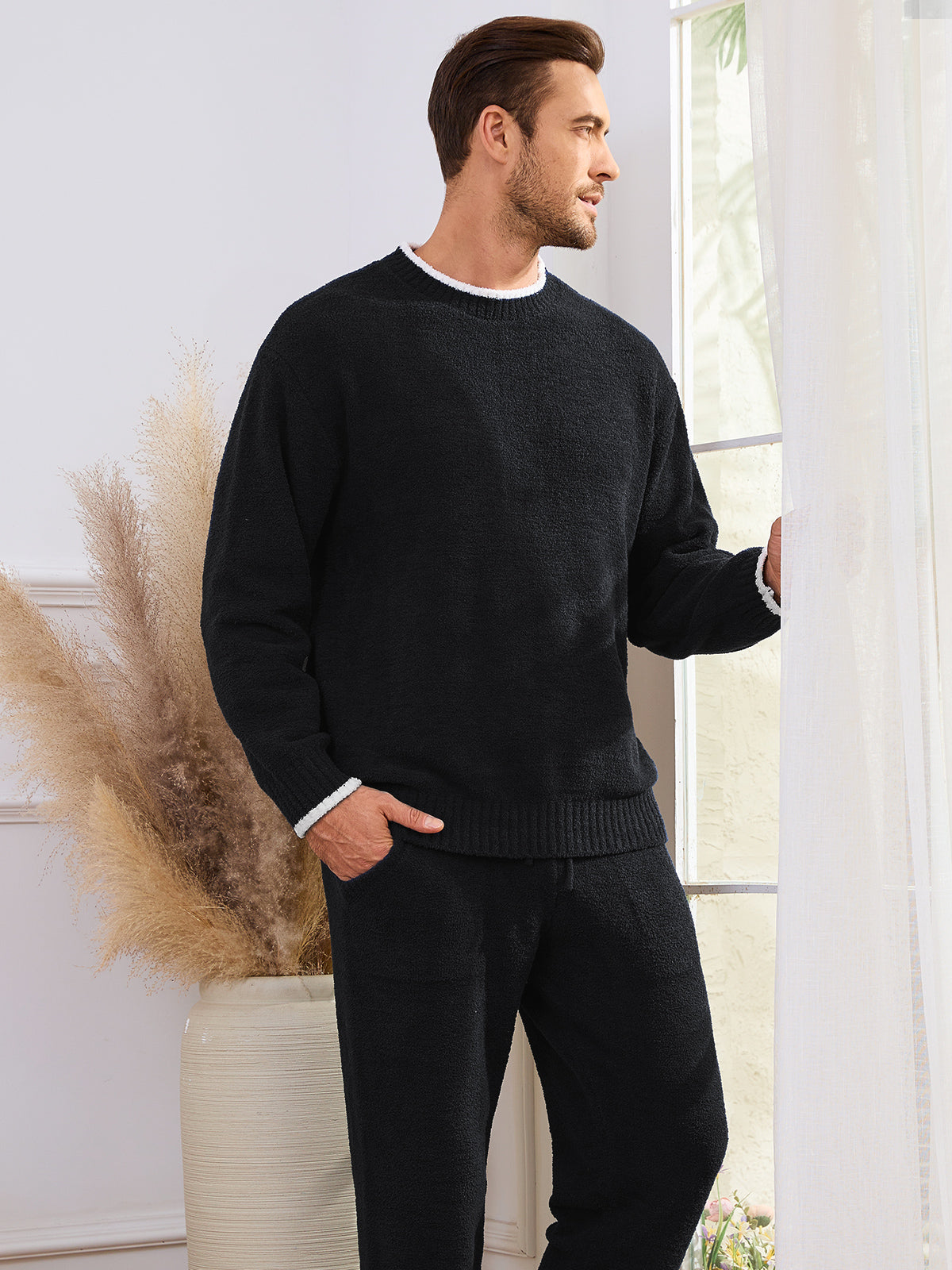Man wearing a black sweater and pants standing in a room with a vase and decorative plant. #color_Black
