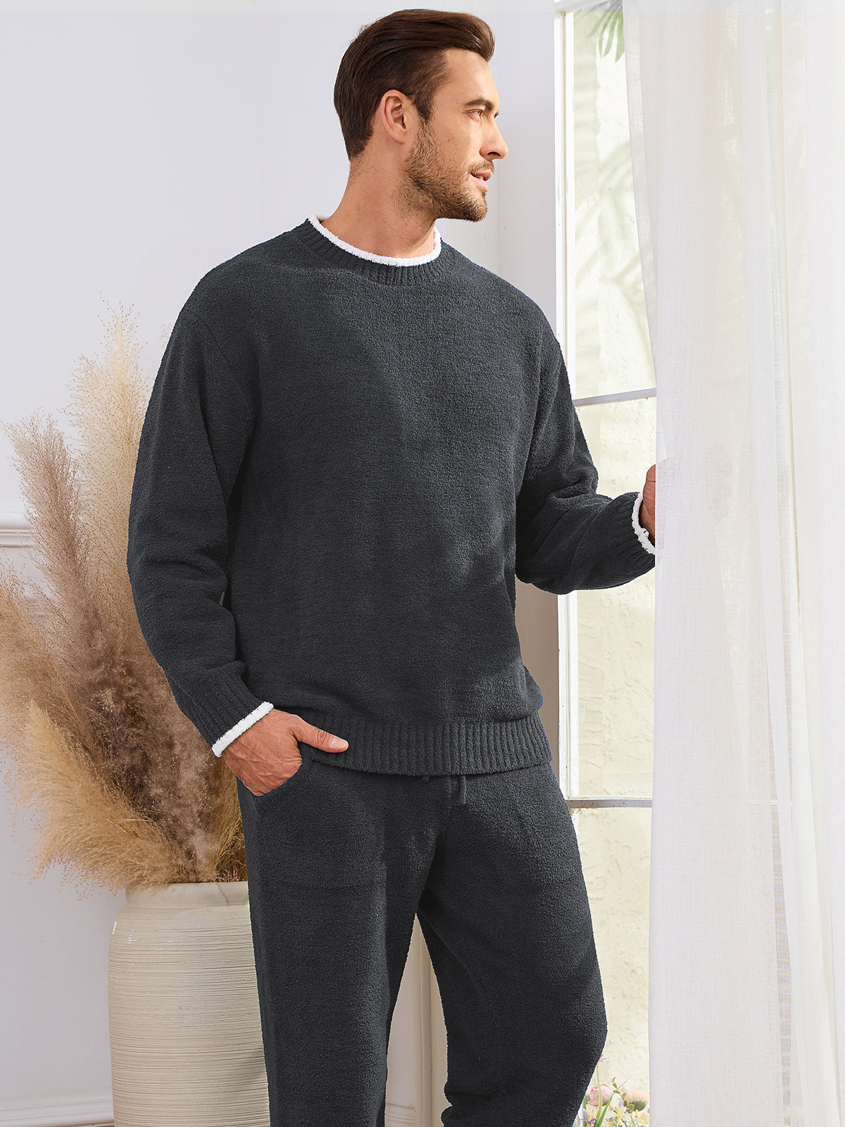 Man wearing a dark gray sweater and pants standing indoors with a window and decorative plant in the background. #color_Dark Grey
