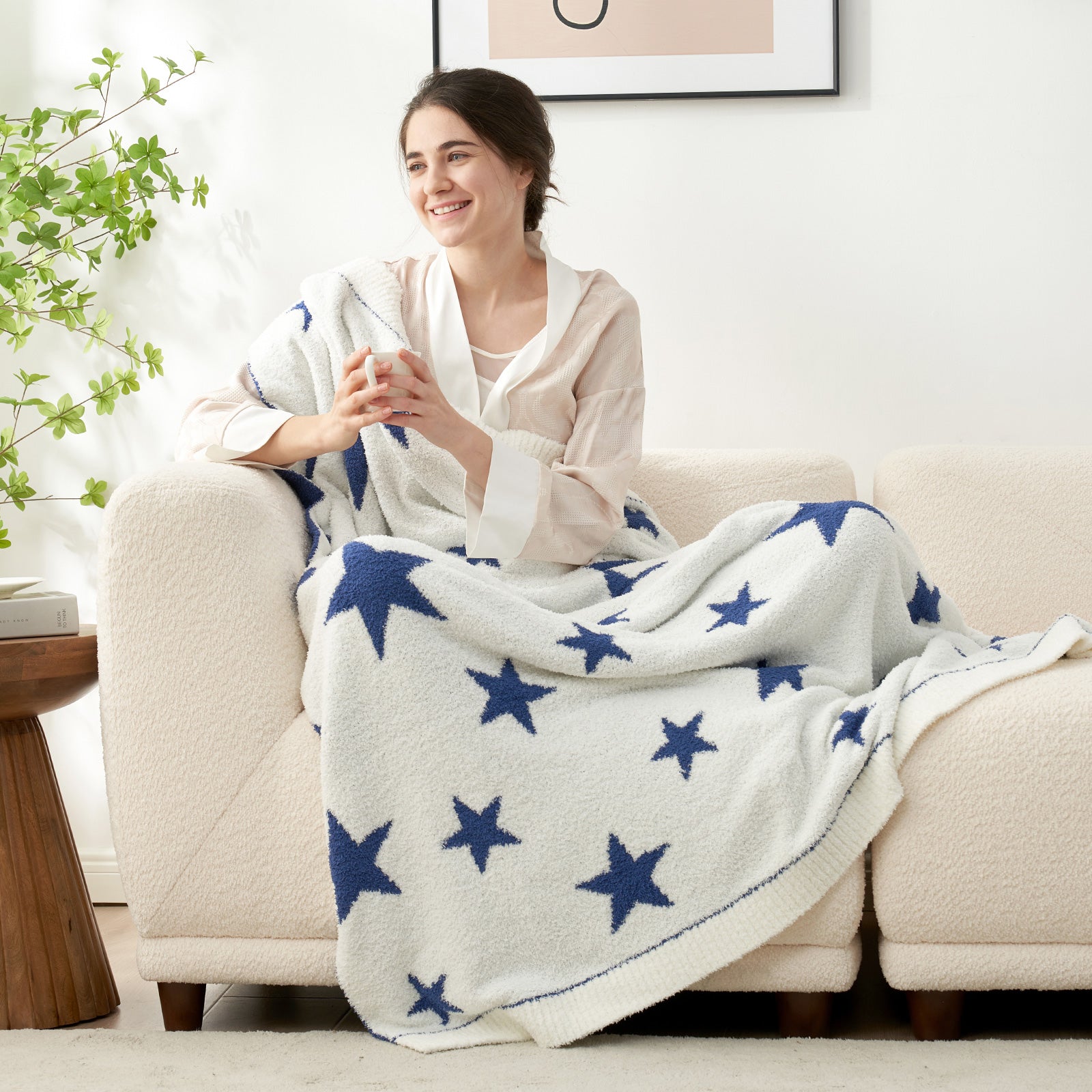 A woman sitting on the beige couch with Snuggle Sacs Reversible Star Feather Yarn Knit Throw Blanket and a mug#color_Royal_Heather_Blue
