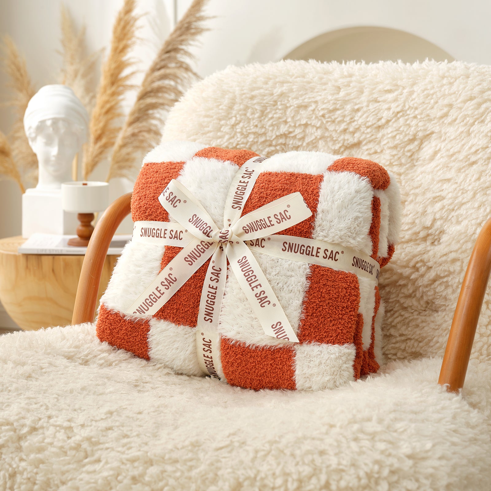 Checkered blanket with 'Snuggle Sac' packaging on a fluffy chair #color_Orange