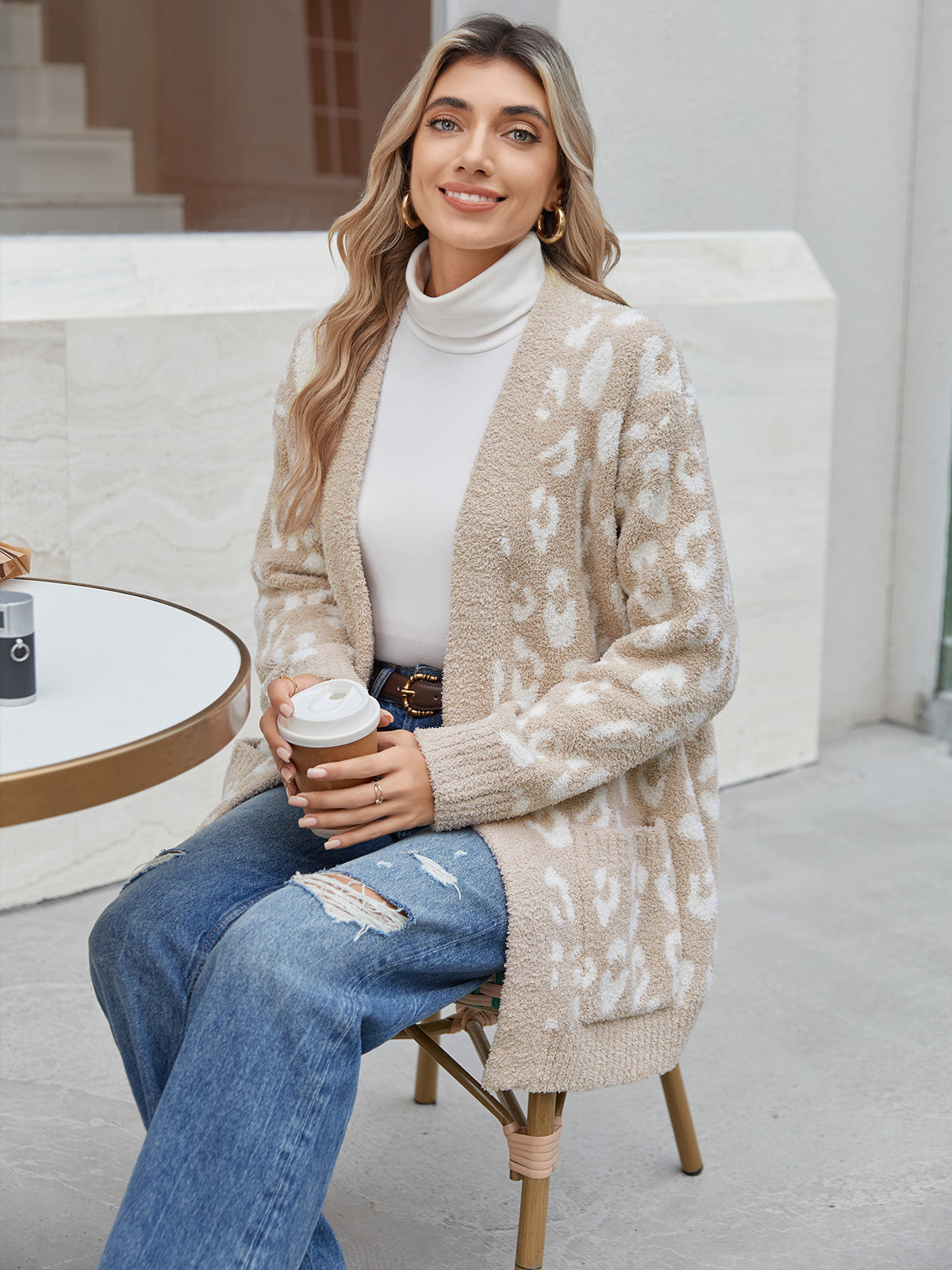 Woman sitting outdoors holding a coffee cup, wearing a patterned cardigan and jeans. #color_Leopard Beige
