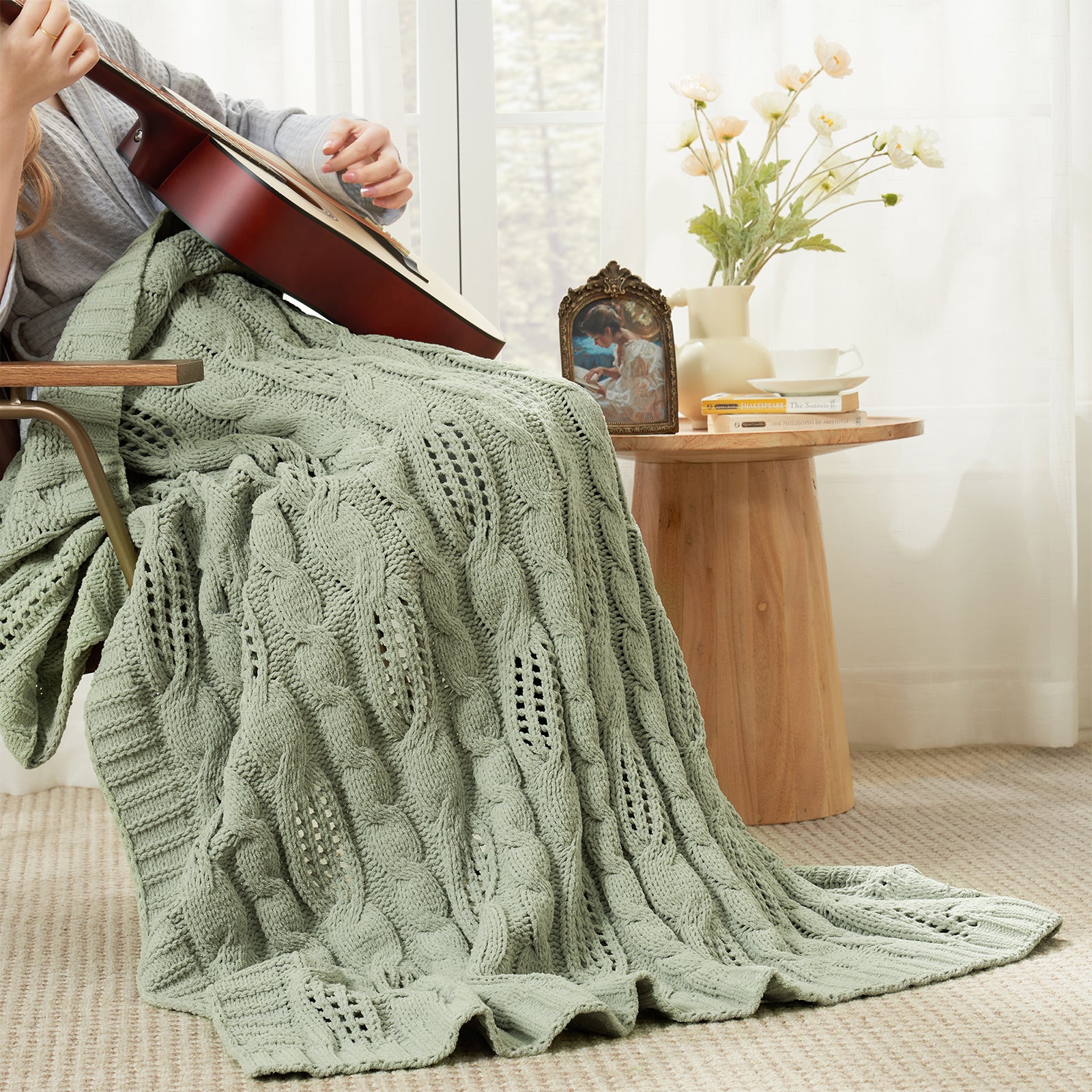 Woman sitting on chair with a Snuggle Sac cable chenille knit throw blanket in Sage Green while playing guitar #color_Sage Green