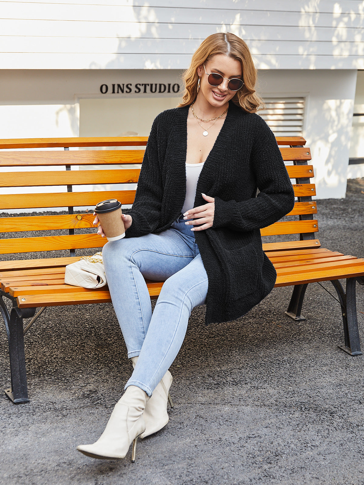 Woman sitting on a bench wearing a black cardigan, light blue jeans, and white boots, holding a coffee cup.#color_Black #color_Black