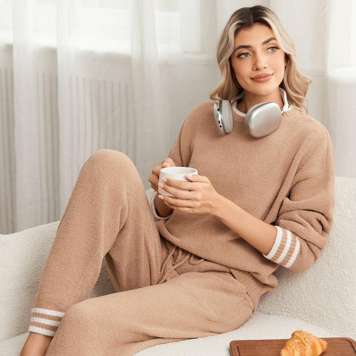 Woman in a beige lounge set sitting on a couch with a coffee cup and pastry. #color_Khaki
