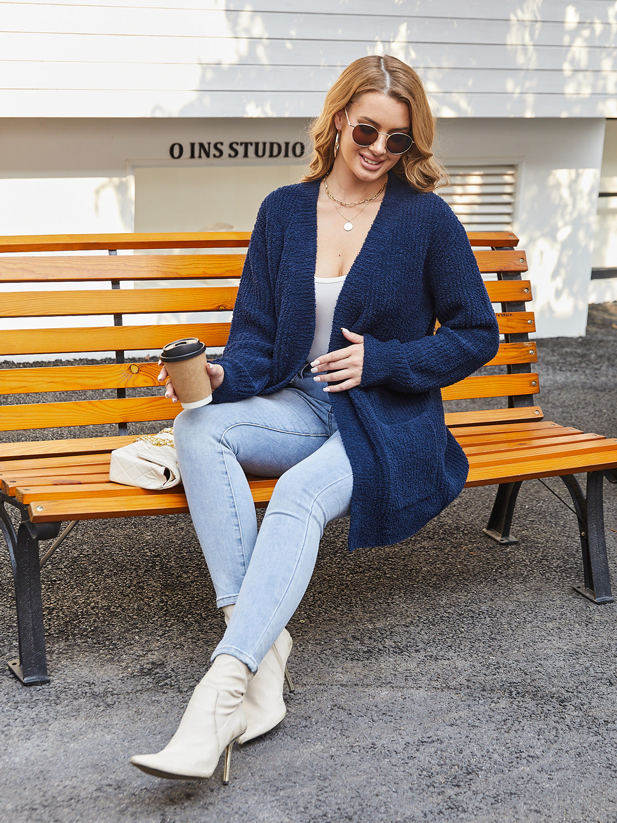 Woman sitting on a bench wearing a blue cardigan, light blue jeans, and white boots, holding a coffee cup. #color_Navy Blue