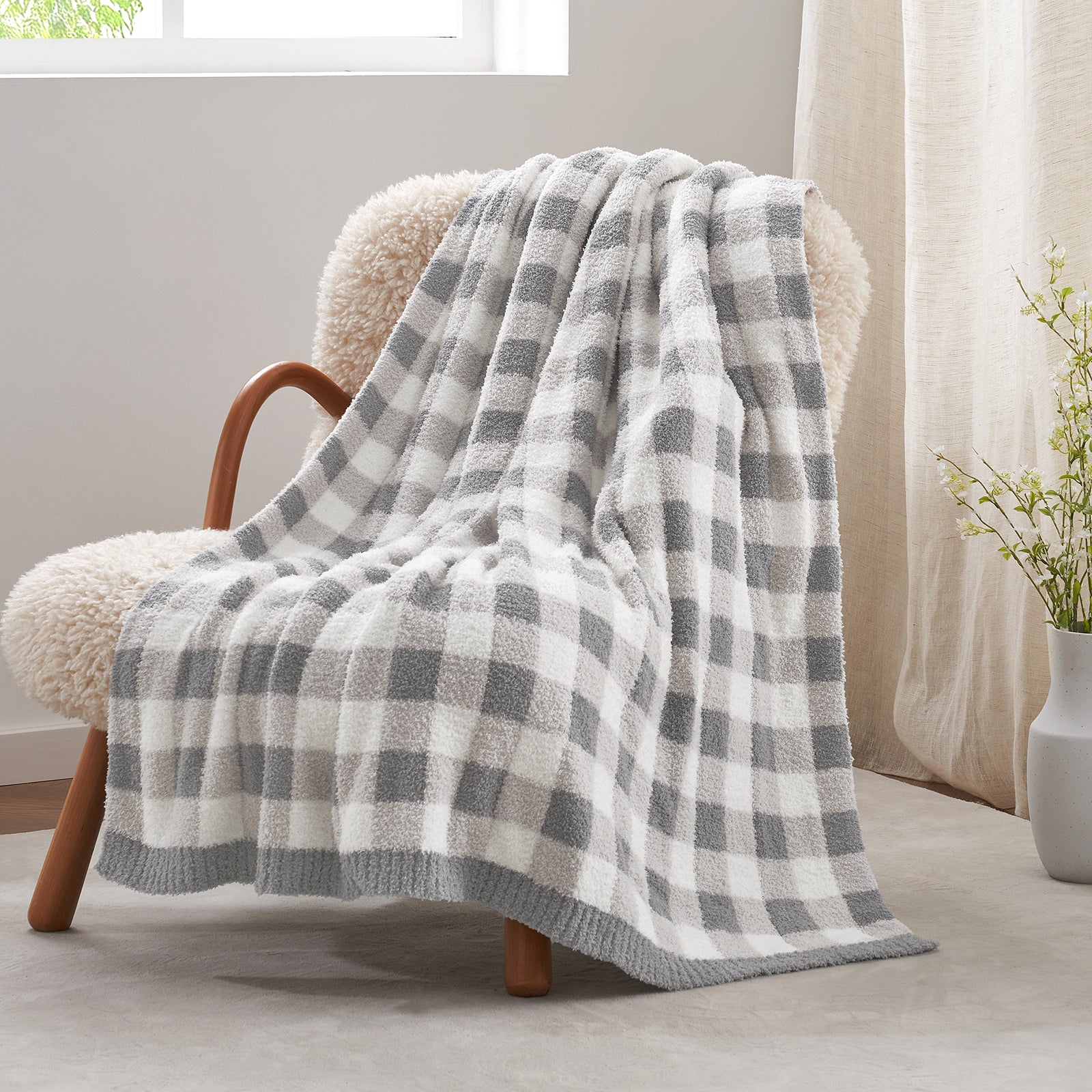 Plaid blanket draped over a chair in a cozy room with a window and plant in the background. #color_Plaid Grey