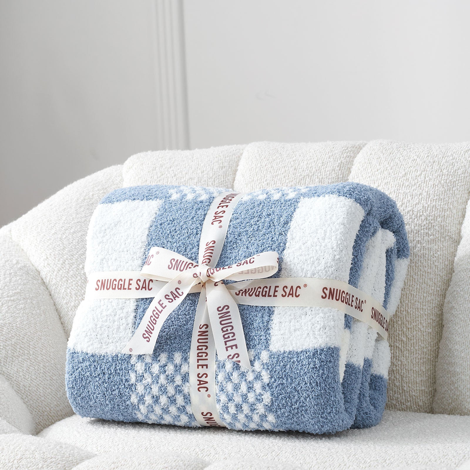 Blue and white checkered blanket with 'Snuggle Sac' label on a light gray couch. #color_Pale Blue