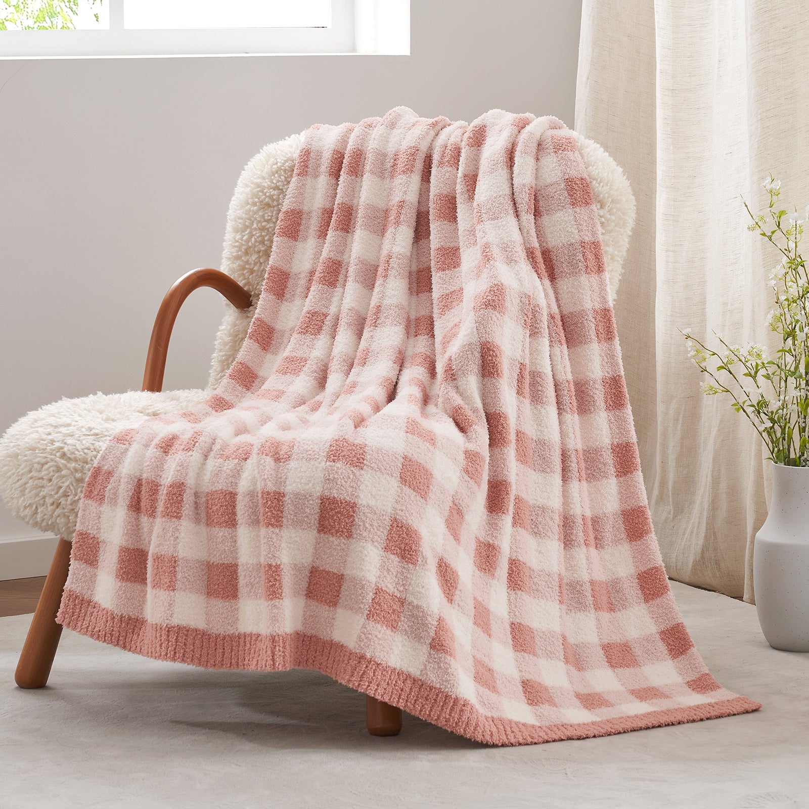 Pink and white checkered blanket draped over a chair in a bright room with a vase of flowers.#color_Plaid Pink