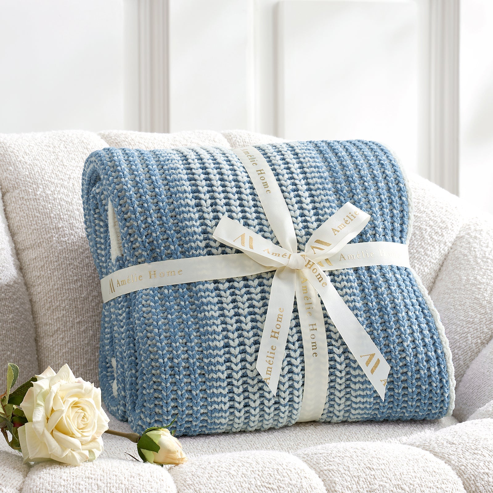 Blue knitted blanket with a white ribbon on a light-colored couch, accompanied by white roses. #color_Two-tone Blue