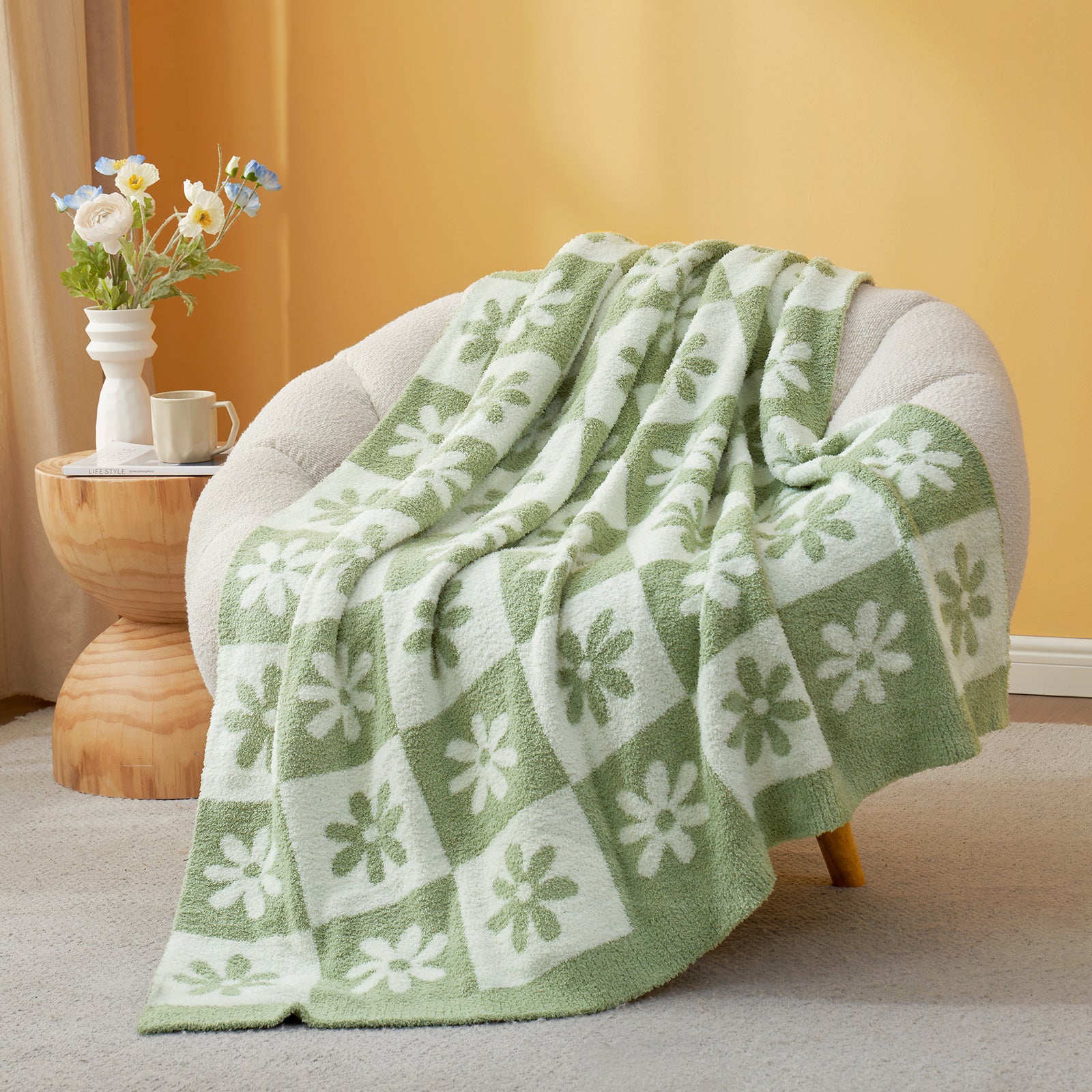 Green and white patterned blanket draped over a chair with a side table and flowers in the background. #color_Sage Green