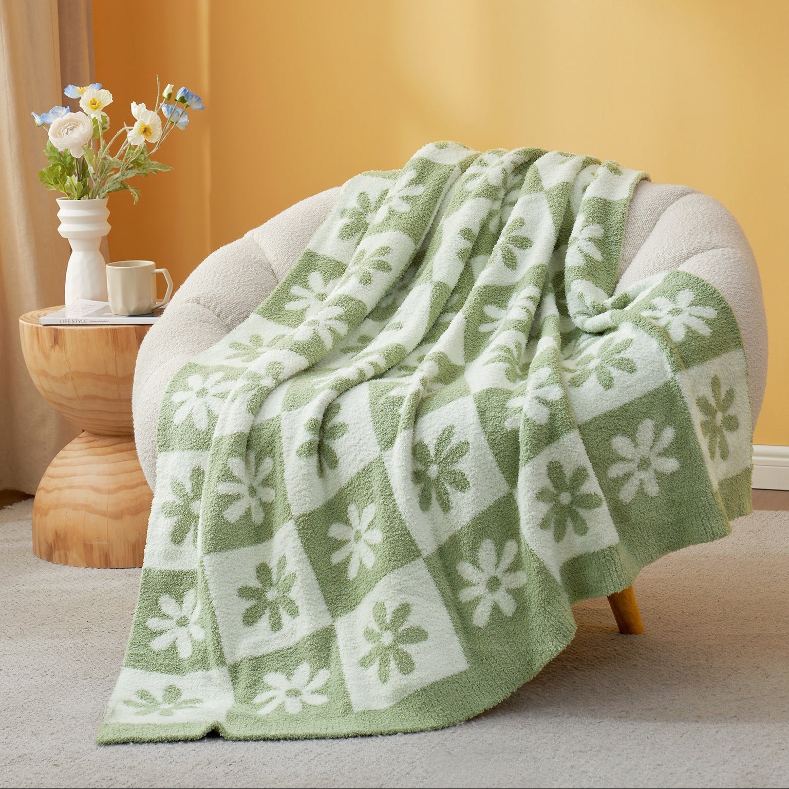 Green and white patterned blanket draped over a chair with a side table and flowers in the background. #color_Sage Green