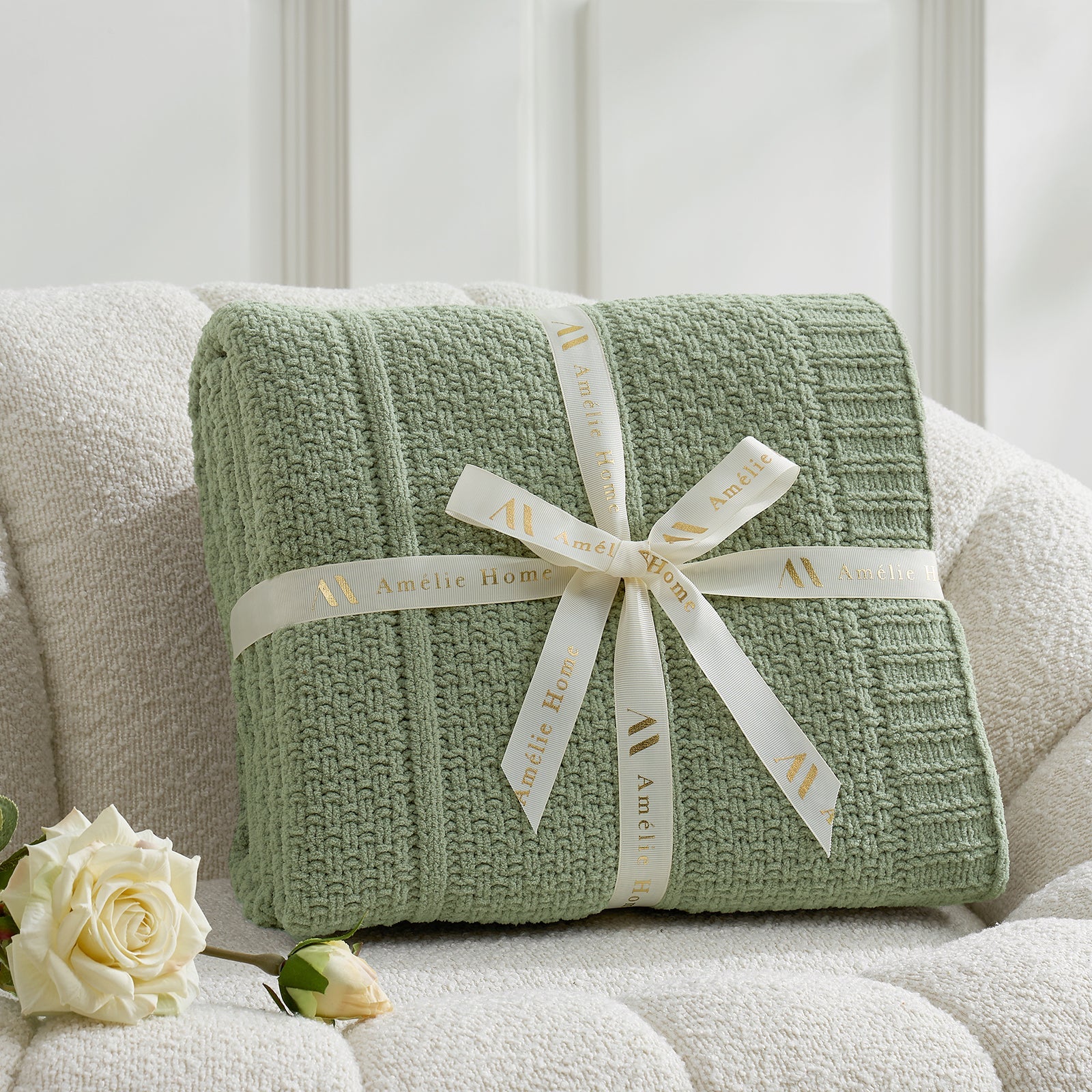 Green textured blanket with a white ribbon on a light-colored couch, accompanied by a white rose. #color_Sage Green