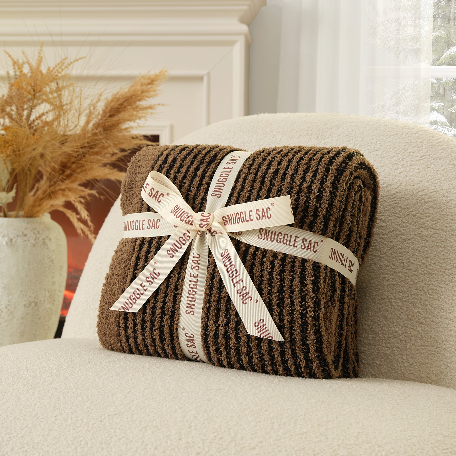 Brown knitted blanket with 'Snuggle Sac' label on a beige armchair. #color_Brown/Black