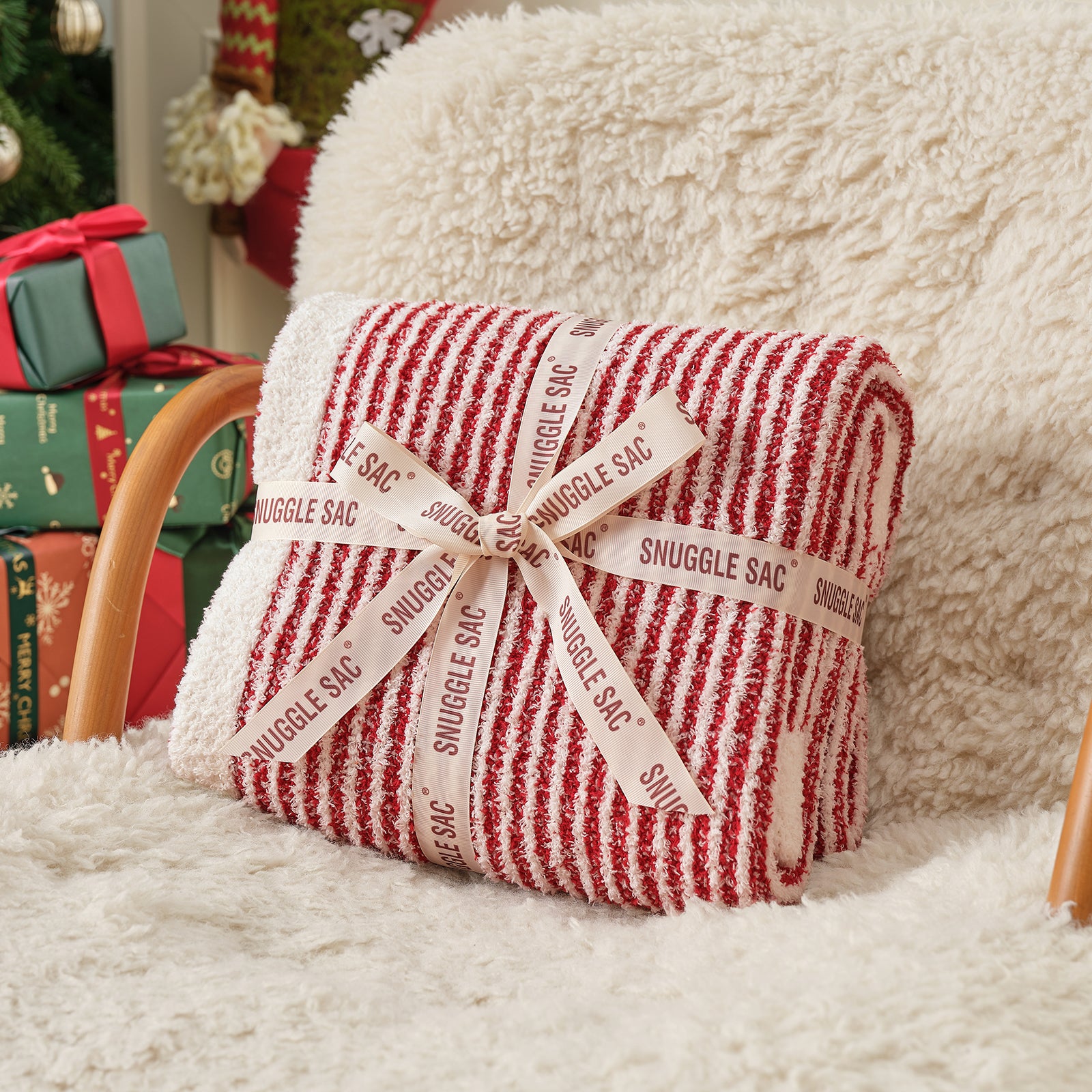 Red and white striped blanket with 'Snuggle Sac' label on a fluffy surface, Christmas decorations in the background. #color_Red/Cream