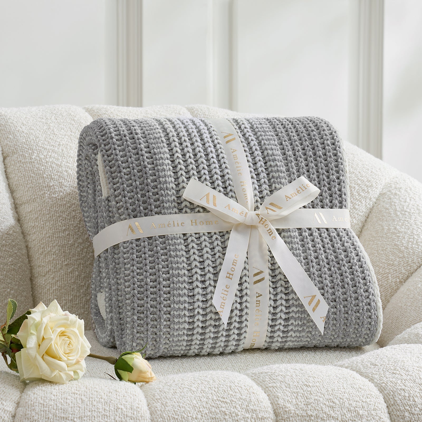 Grey knitted blanket with a white ribbon on a beige sofa with white roses. #color_Two-tone Grey