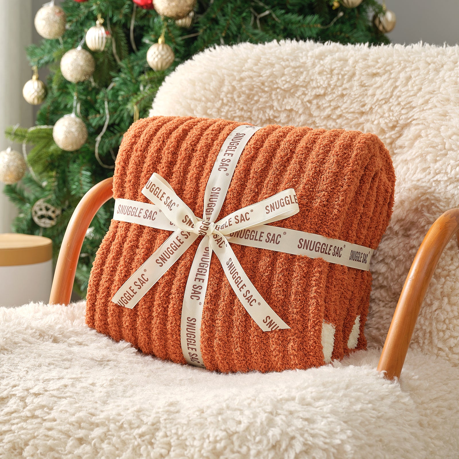 Orange snuggle sac with a decorative bow on a fluffy chair with a Christmas tree in the background #color_Burnt Orange