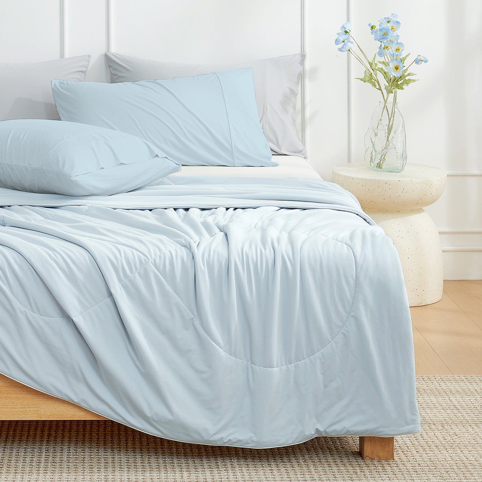 Blue bedding set on a bed in a bedroom with a vase of flowers on a nightstand.#color_Blue