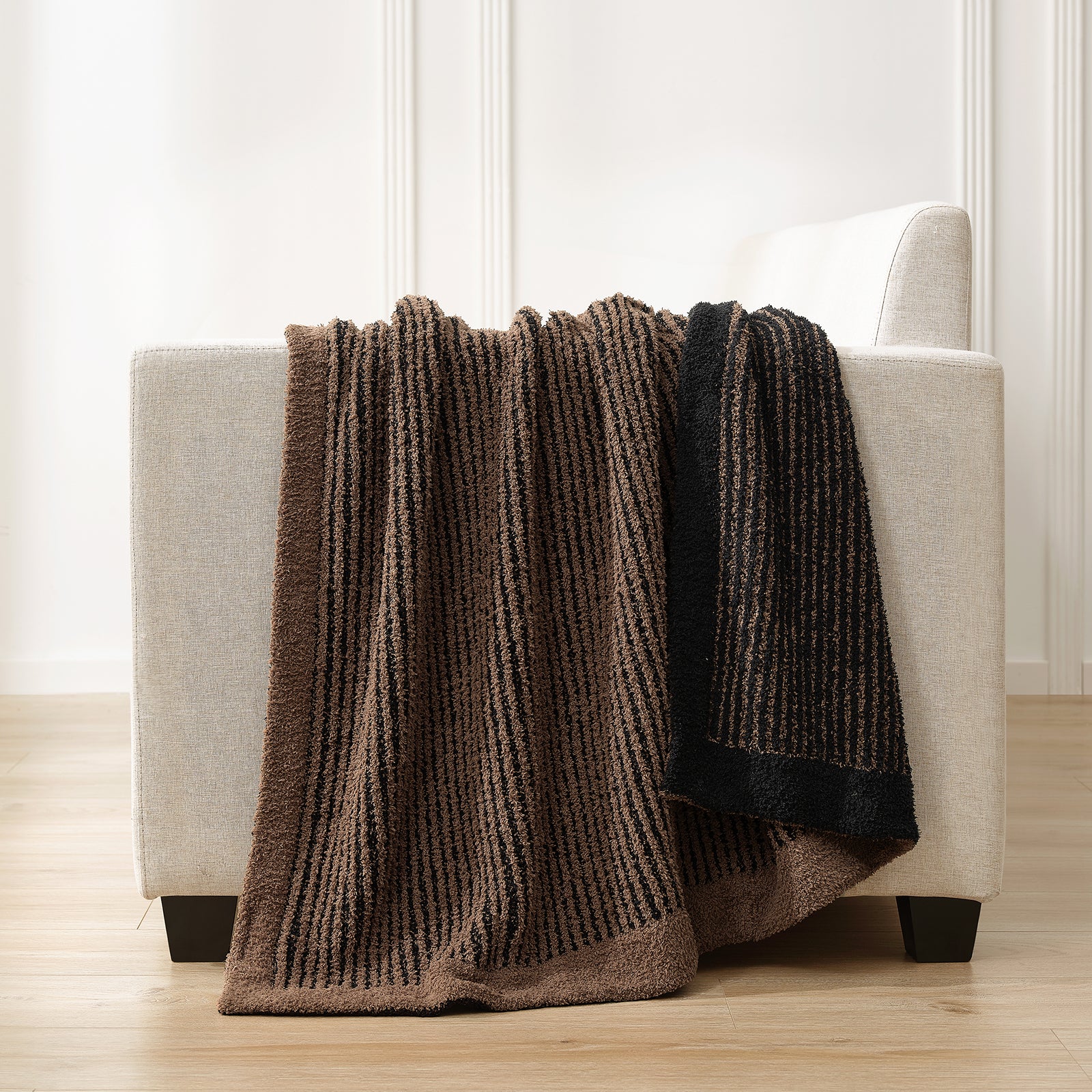 Brown textured blanket draped over a white armchair in a room with light wood flooring and white walls. #color_Brown/Black