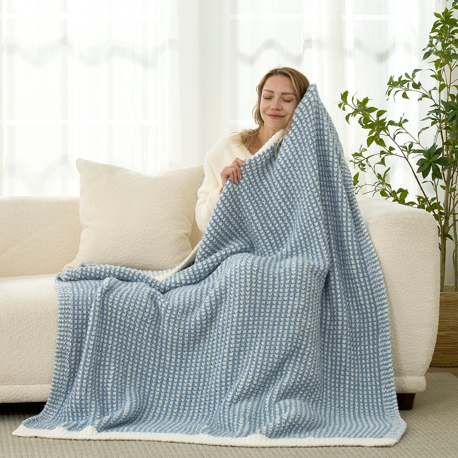 One Woman sitting on a couch under a large blue blanket in a bright room. #color_Cream/Blue