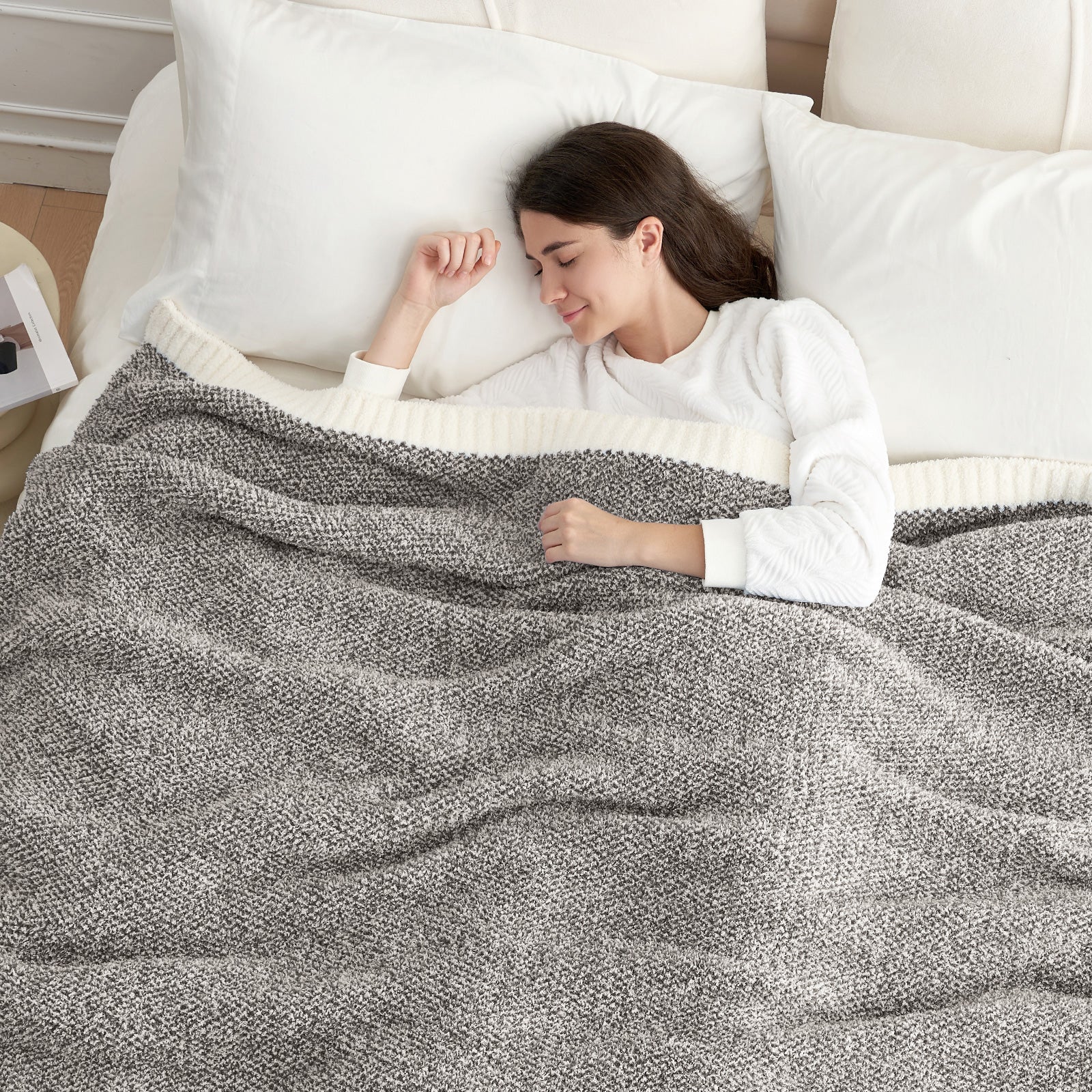 Woman lying on a bed under a gray textured blanket, wearing a white sweater.#color_Dark Grey