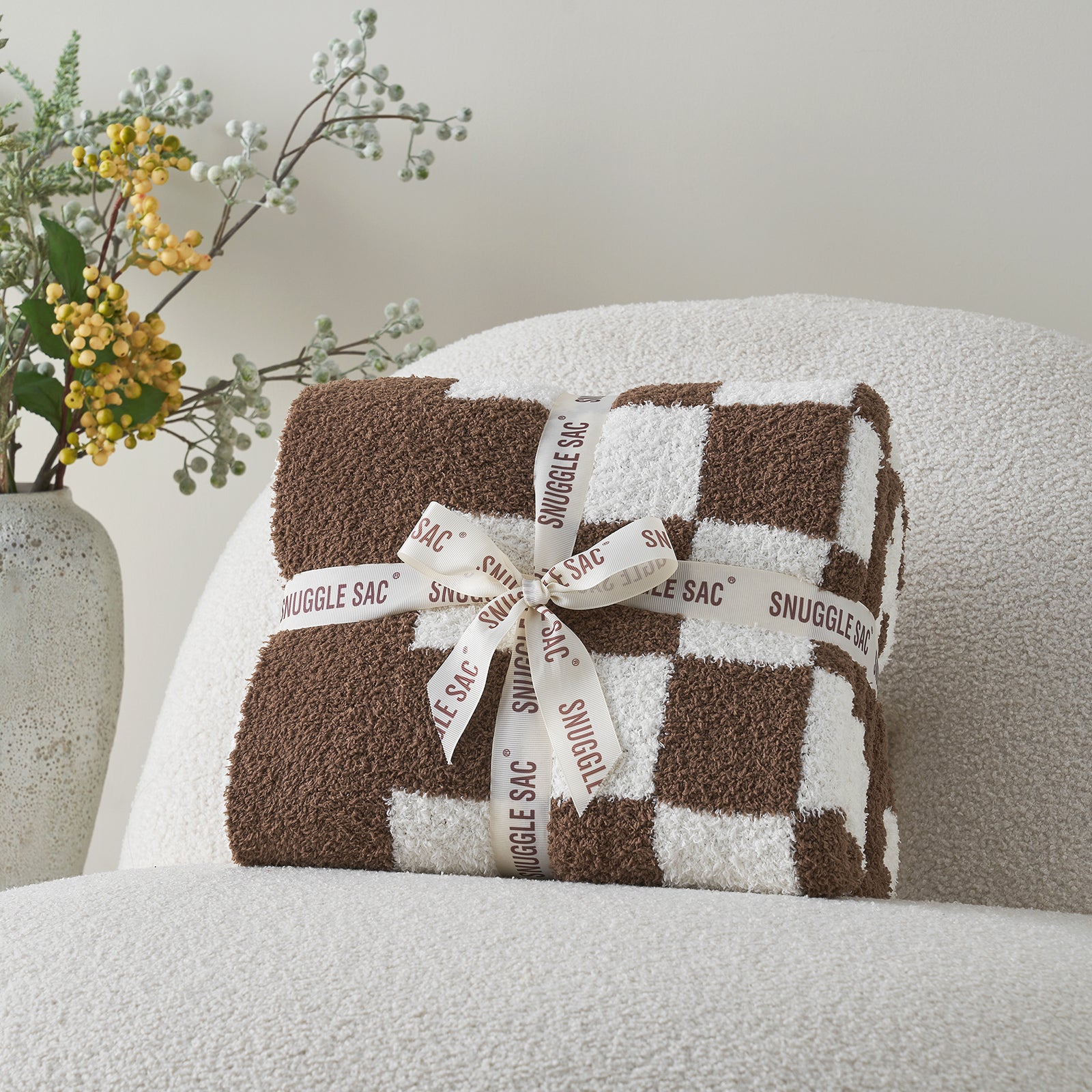 Checkered blanket with a bow labeled 'Snuggle Sac' on a chair next to a vase with flowers. #color_Checkered Brown