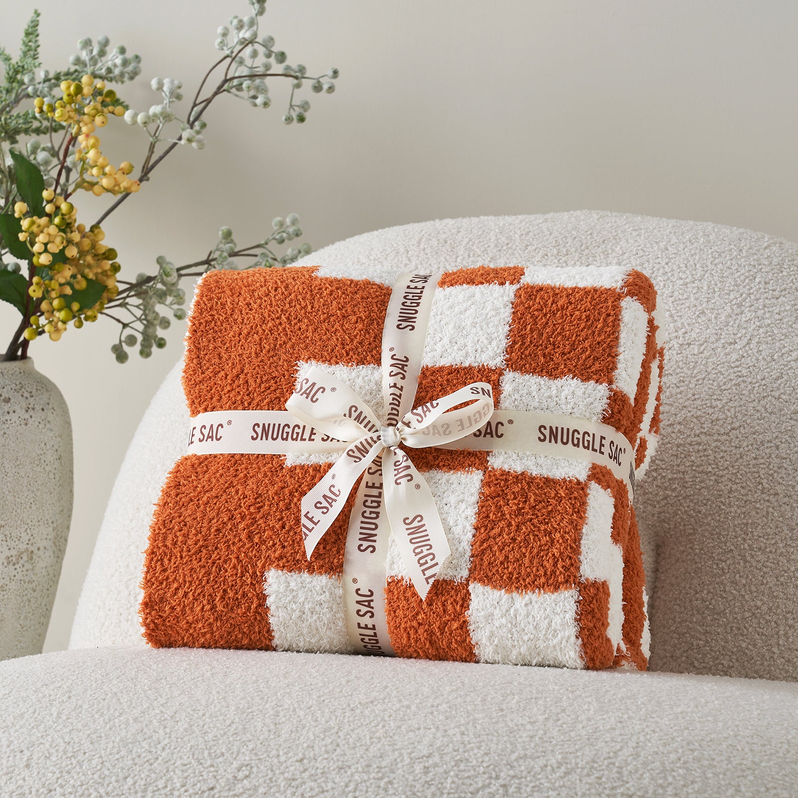 Orange and white checkered blanket with 'Snuggle Sac' ribbon on a chair. #color_Checkered Burnt Orange