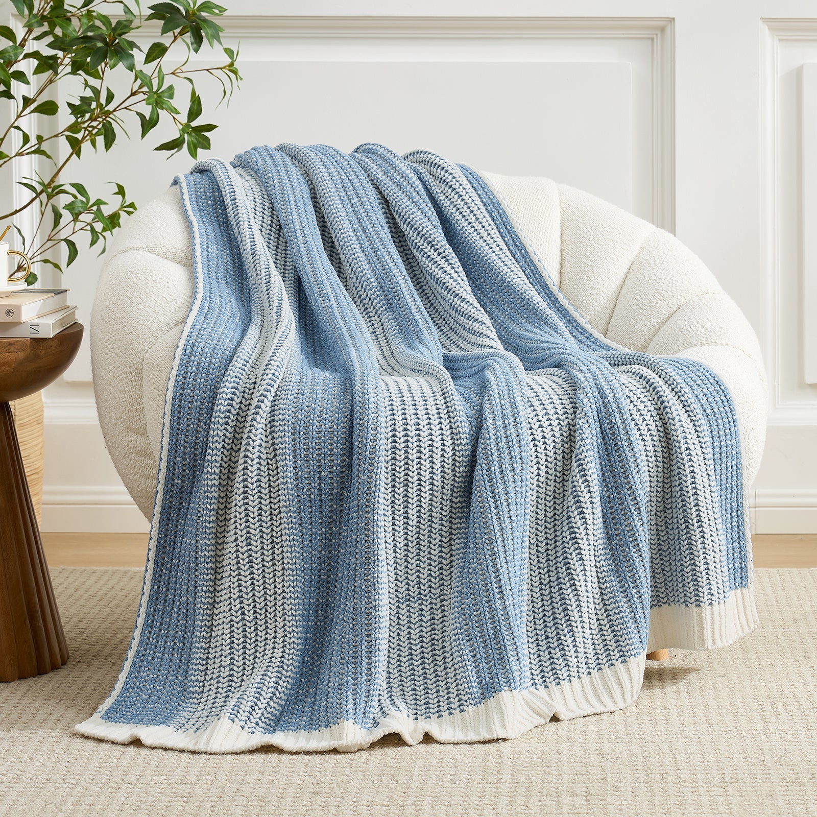 Blue knitted blanket draped over a white armchair in a room with a plant and side table. #color_Two-tone Blue