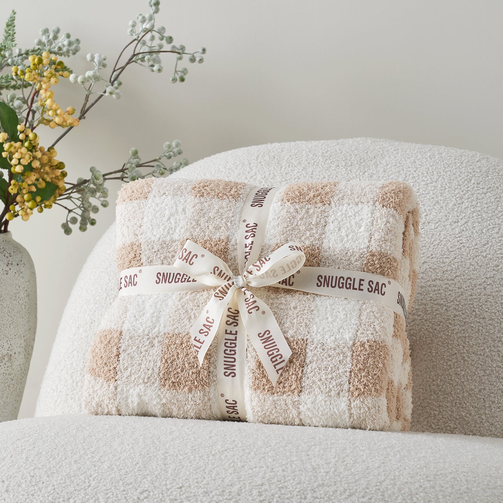Folded blanket with 'Snuggle Sac' label on a chair next to a vase with flowers. #color_Plaid Beige