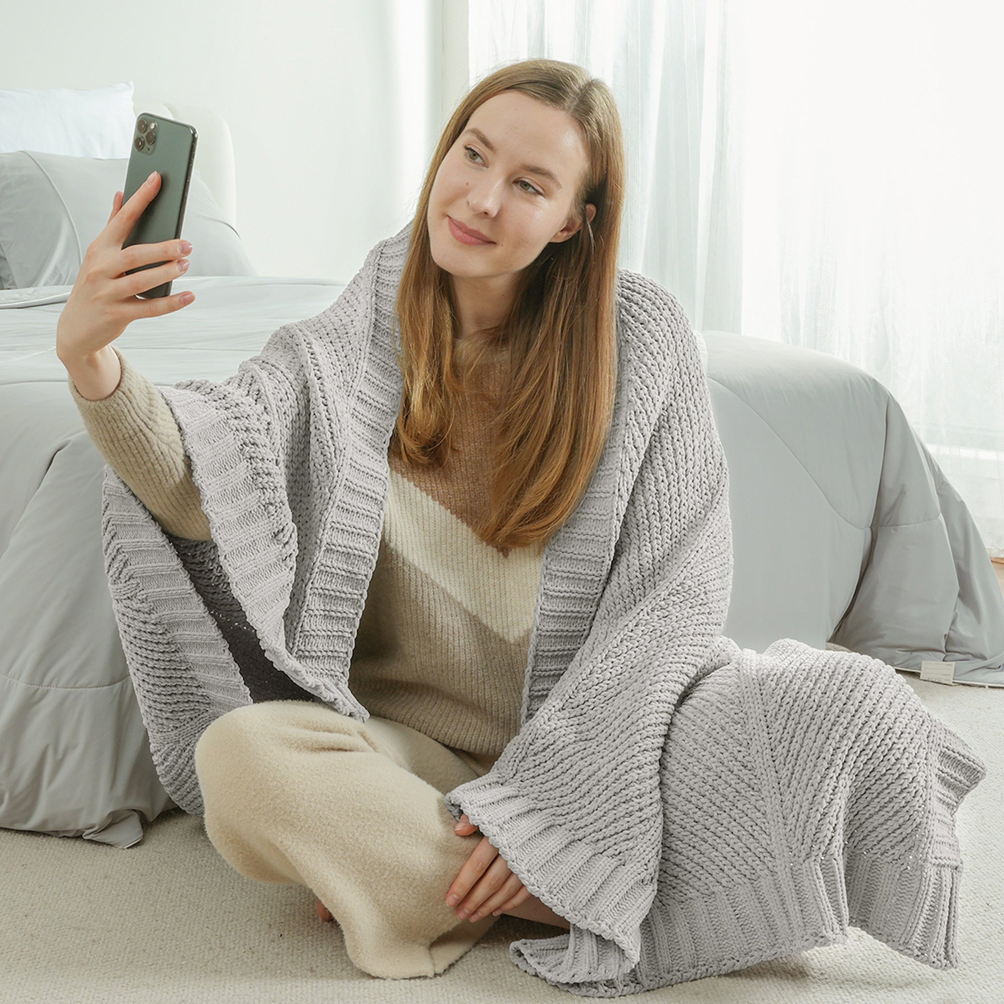 Woman sitting on the floor wrapped in a chenille knit throw blanket in Light Grey taking a selfie #color_Light Grey
