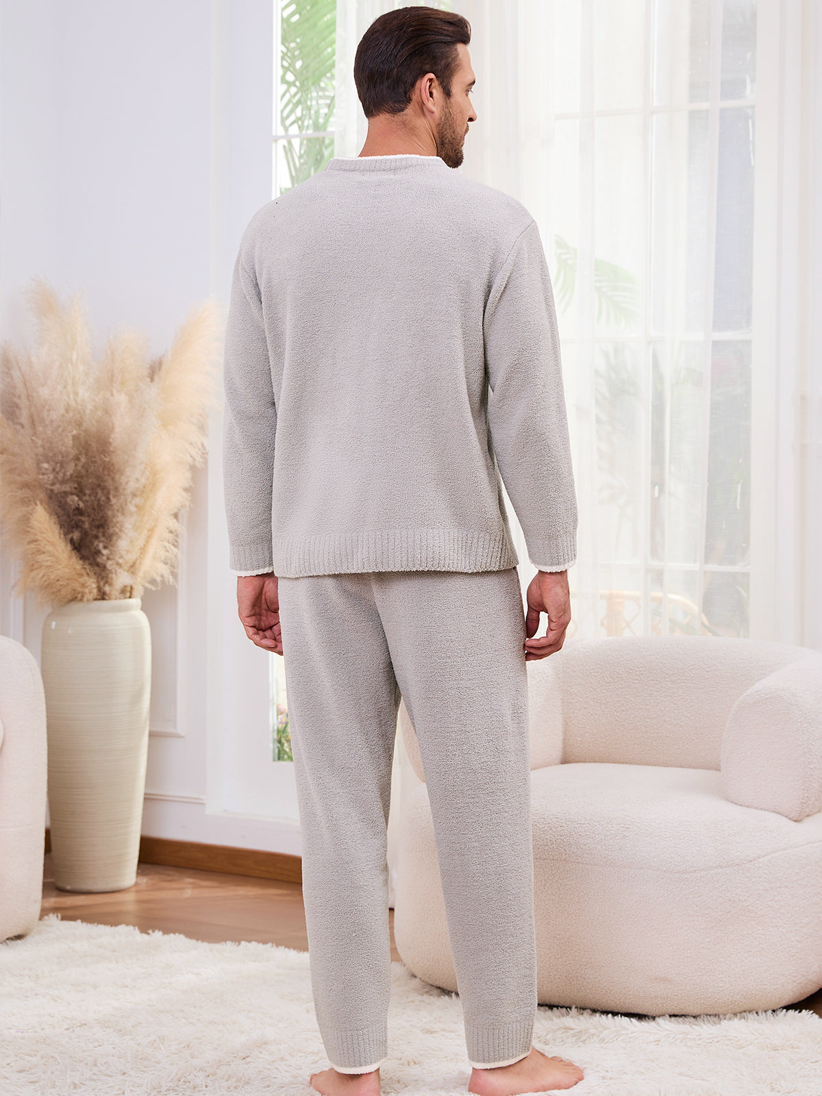Man wearing a gray pajama set standing in a bright living room.  #color_Light Grey