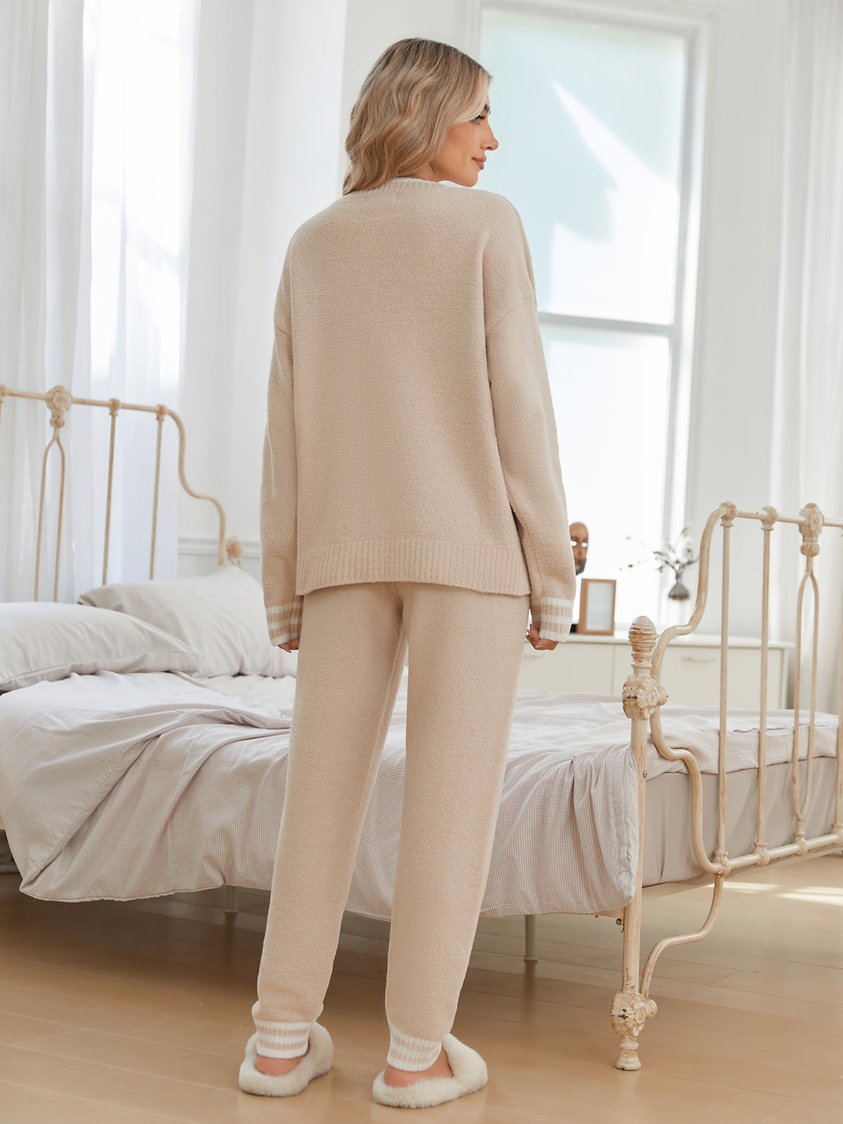 Woman in beige lounge set standing in a bedroom with white curtains and a bed. #color_Beige