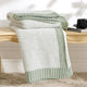 Snuggle Sac knitted green and white blanket on a wooden bench with a cup and book in the background. #color_Sage Green
