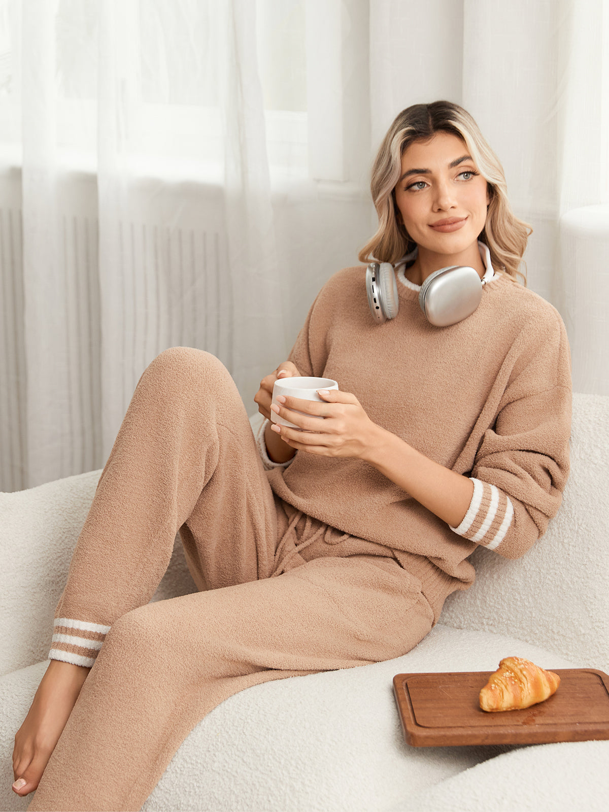 Woman in a beige lounge set sitting on a couch with a coffee cup and pastry. #color_Khaki