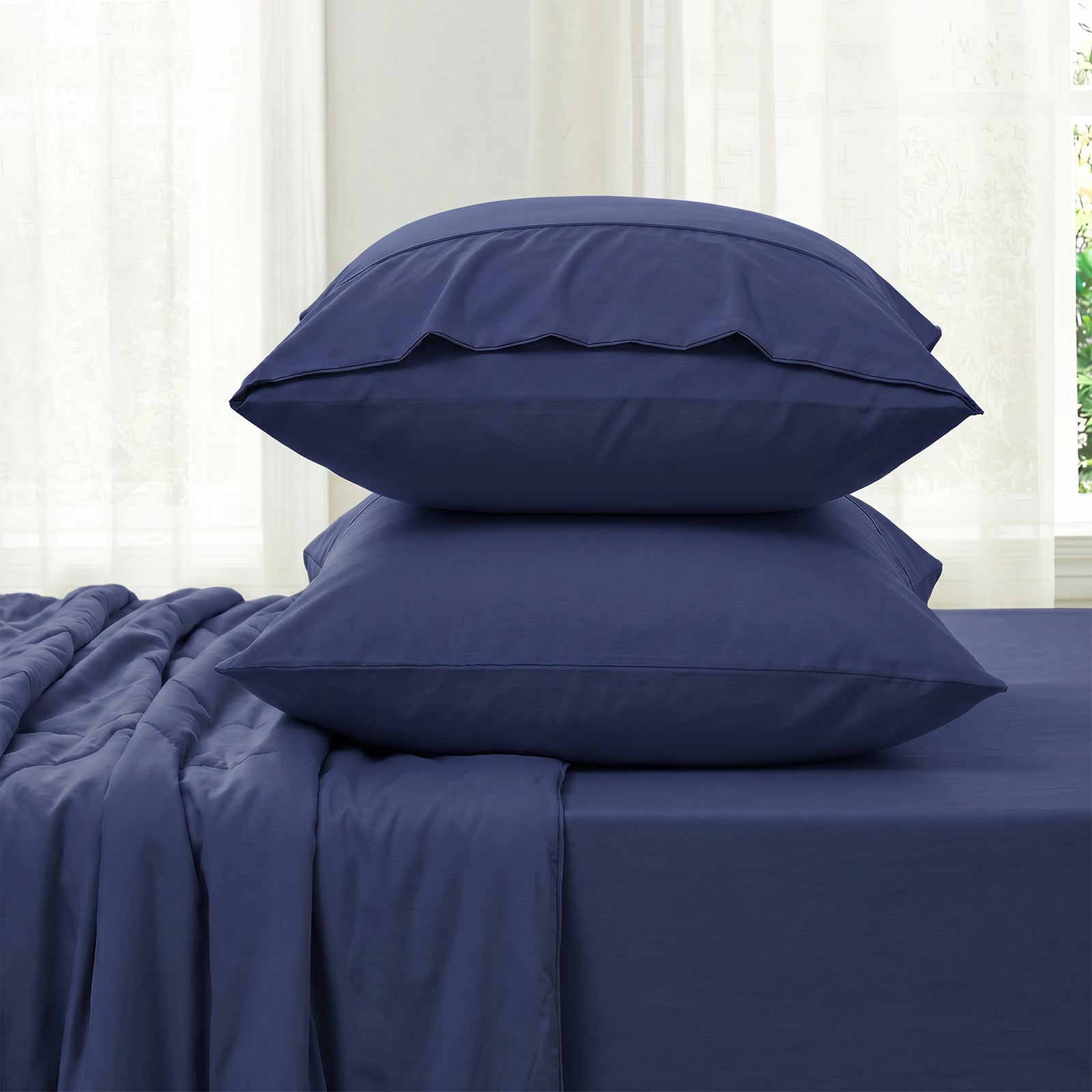 Blue bedding set with two pillowcases and fitted sheet against a white curtain background in side view.#color_Dark Blue