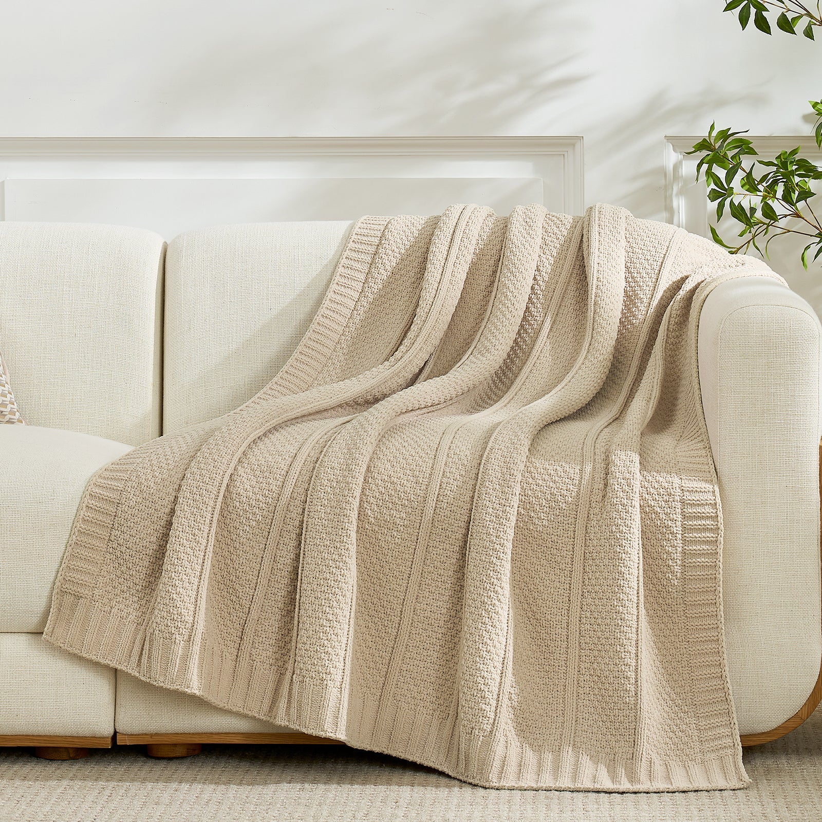Snuggle Sac beige knitted blanket draped over a beige sofa in a bright room with a plant. #color_Beige