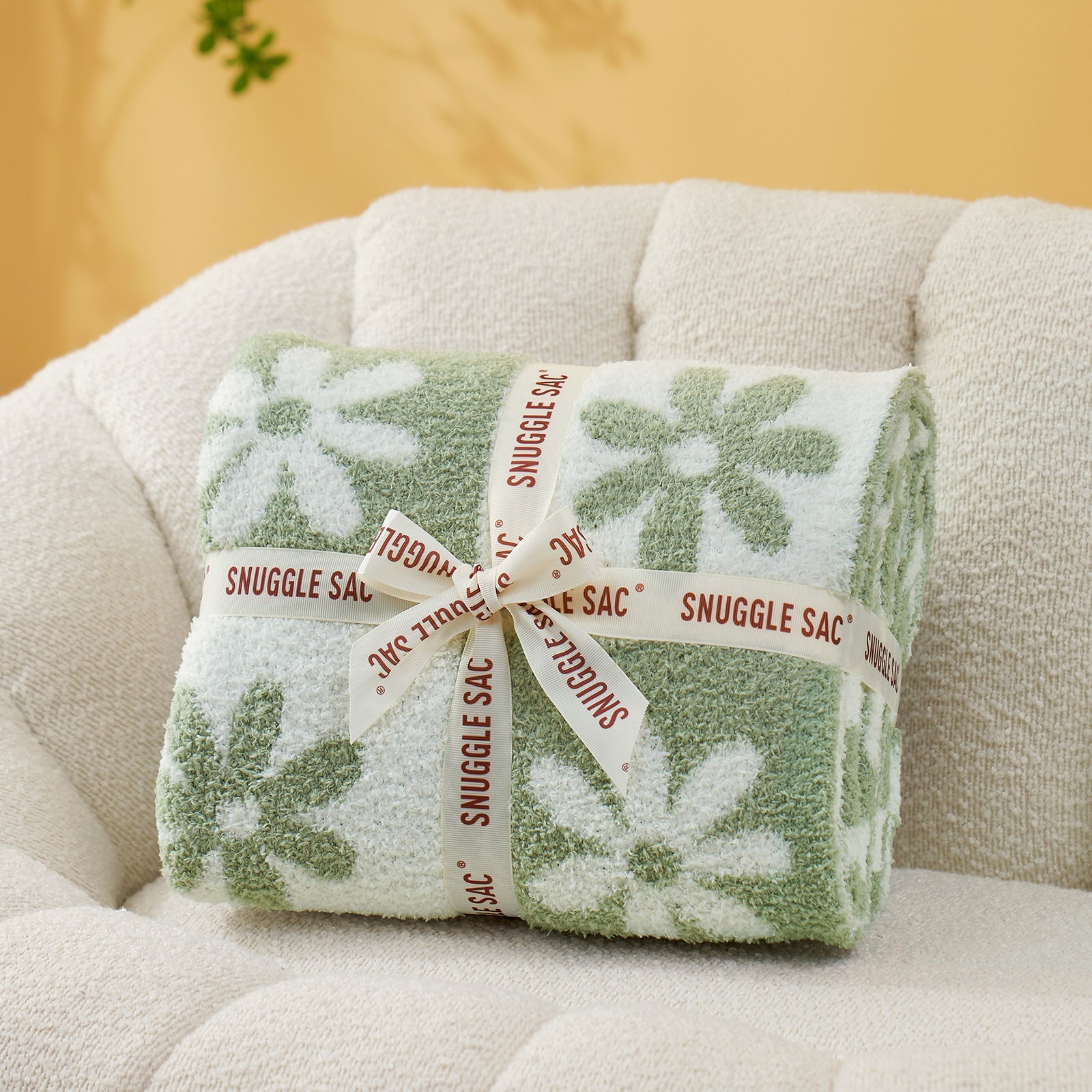 Folded green and white floral blanket with 'Snuggle Sac' label on a beige couch.  #color_Sage Green