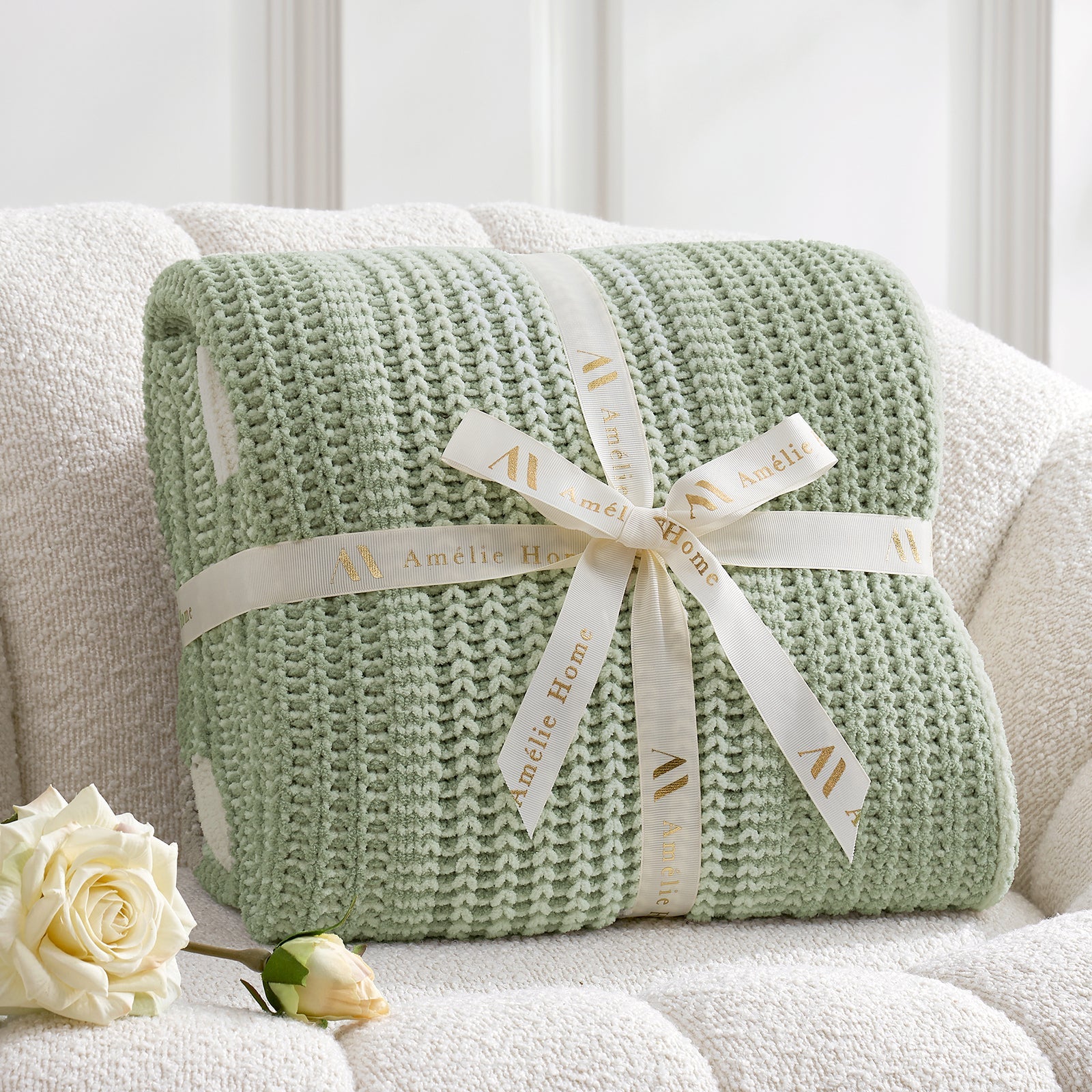 Green knitted blanket with a white ribbon on a white couch, accompanied by a white rose. #color_Two-tone Green